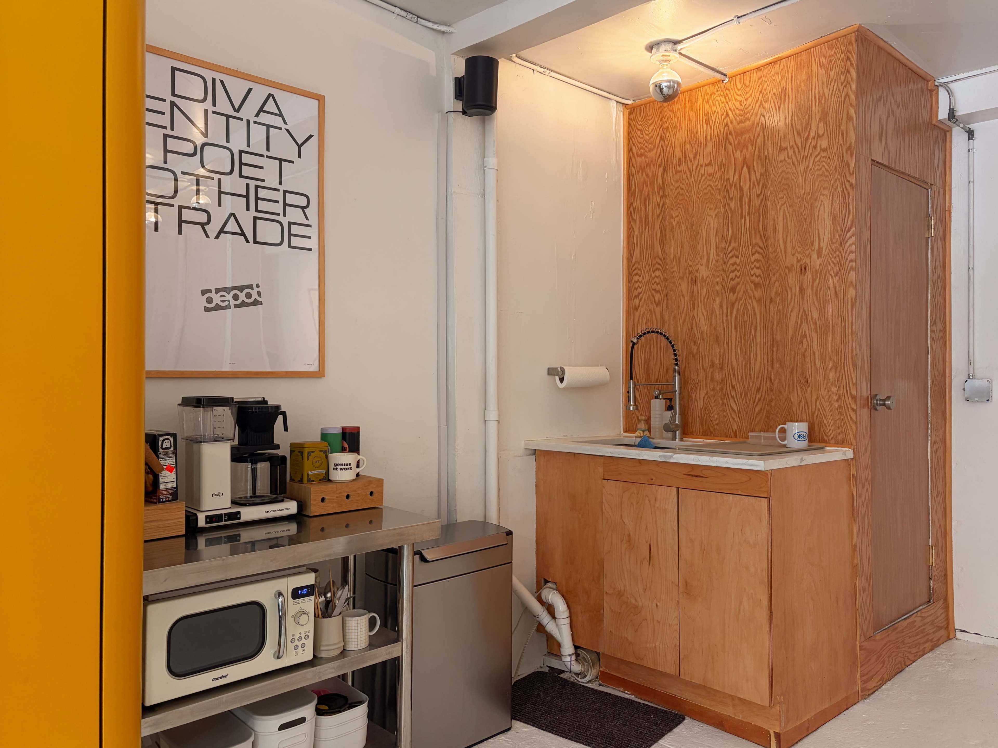 The image shows a small kitchen area with a wooden cabinet, a sink, and various kitchen appliances, including a coffee maker and microwave, set against a white wall with a poster.