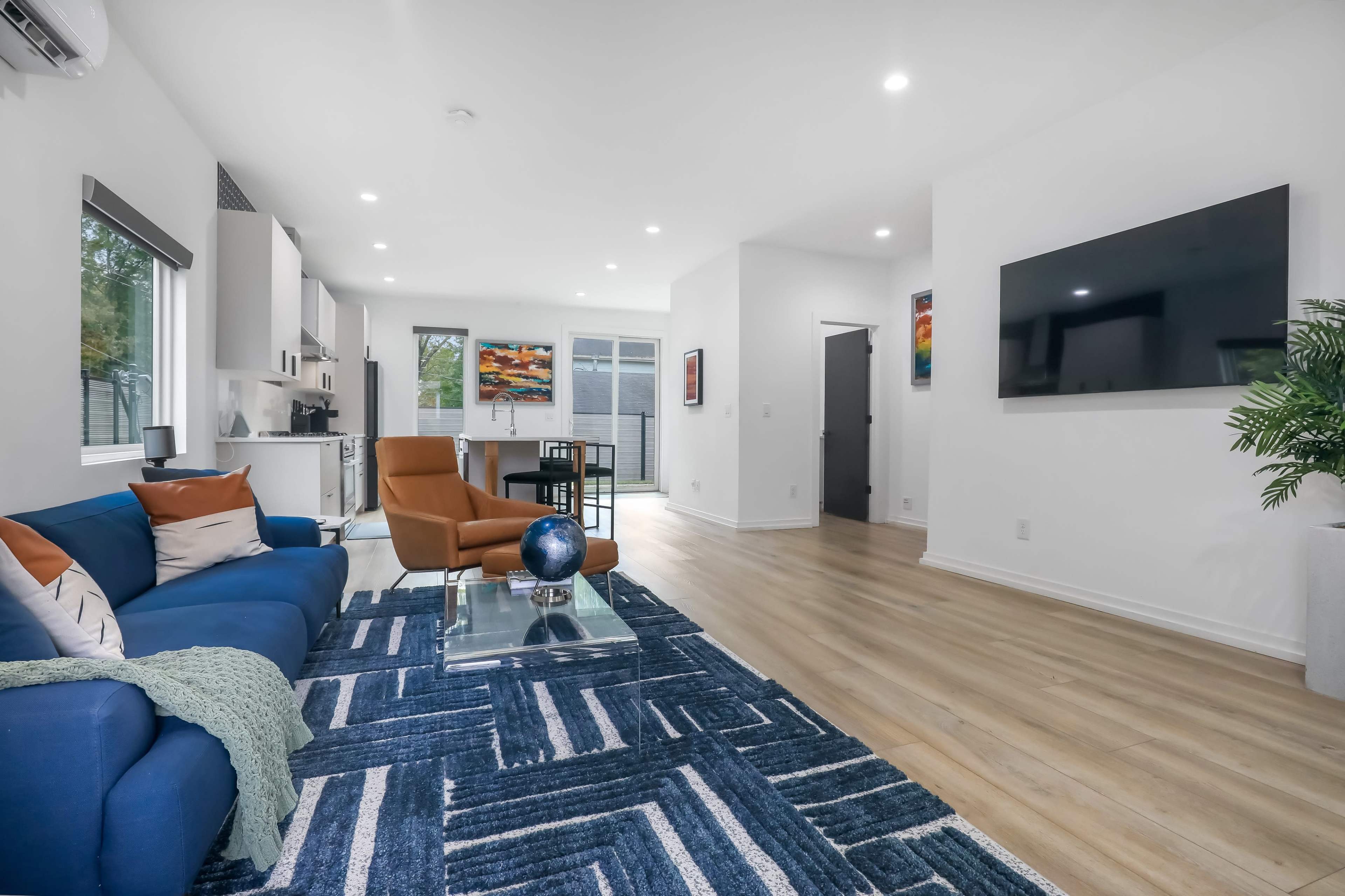 A modern living space with a blue sofa, a glass coffee table, and a large wall-mounted television, complemented by hardwood flooring and natural light from large windows.