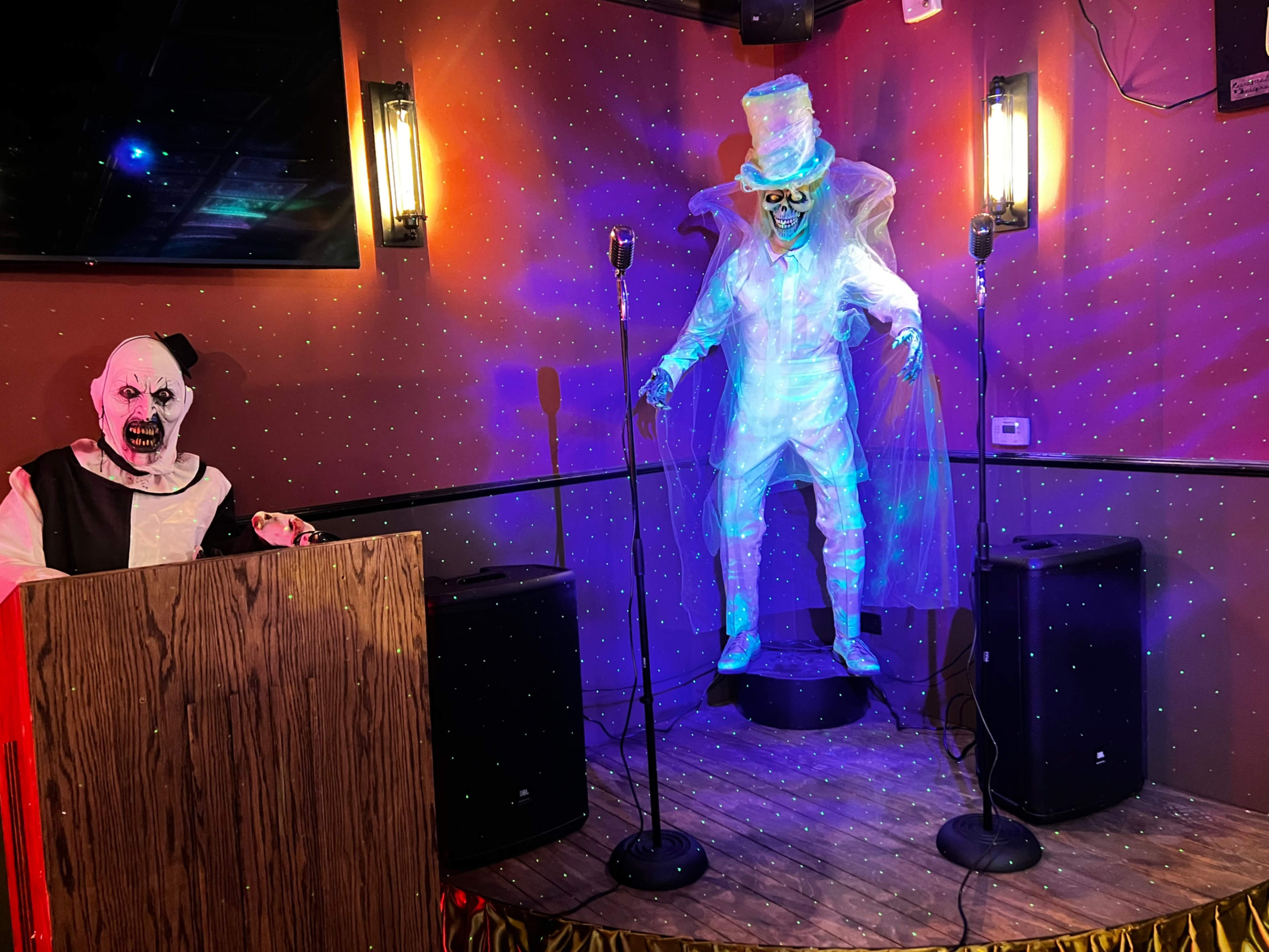 A clown stands behind a podium next to a glowing ghost figure on a stage with microphones.