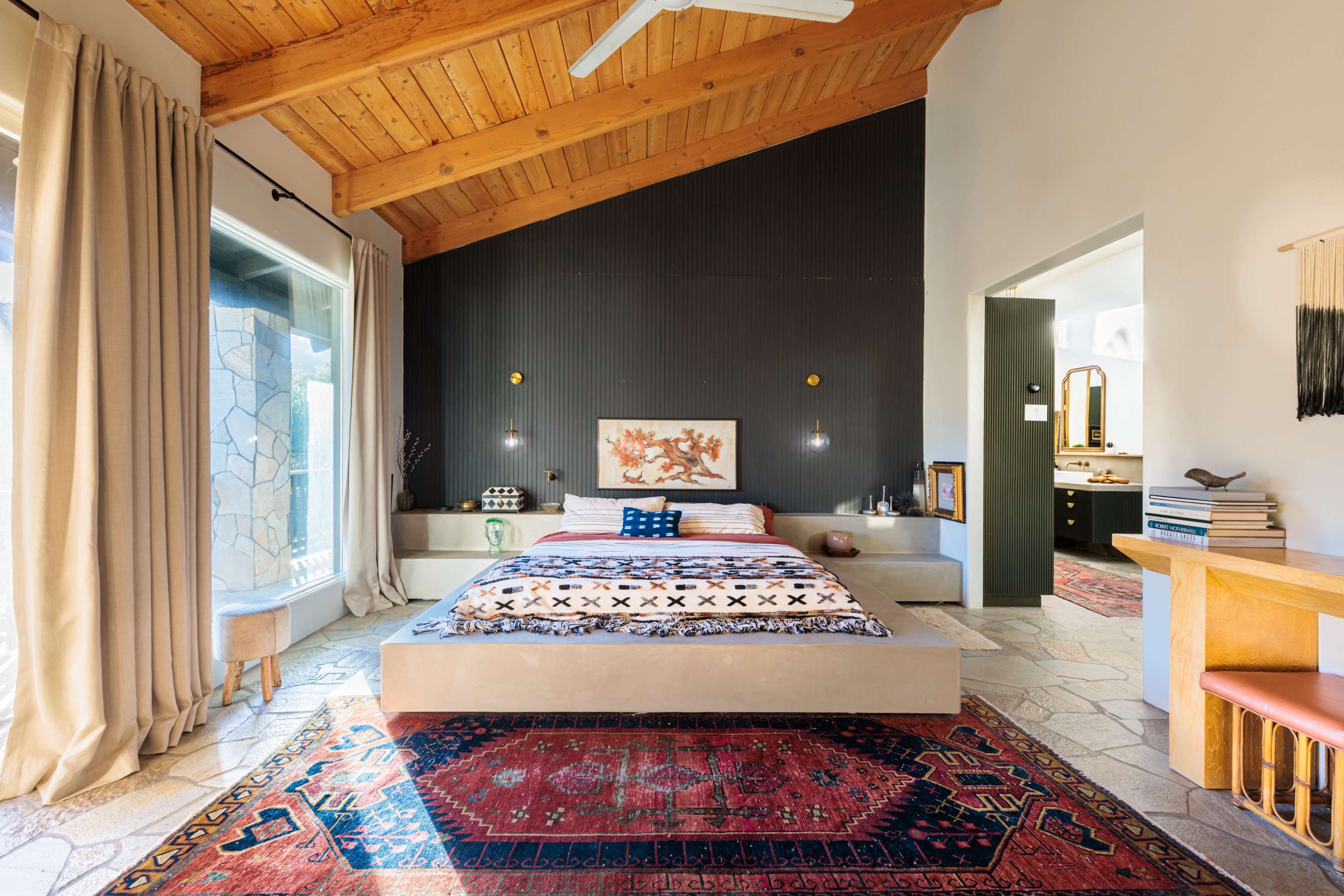The image shows a modern bedroom with a raised bed on a platform, accented by a red area rug and wooden ceiling beams.