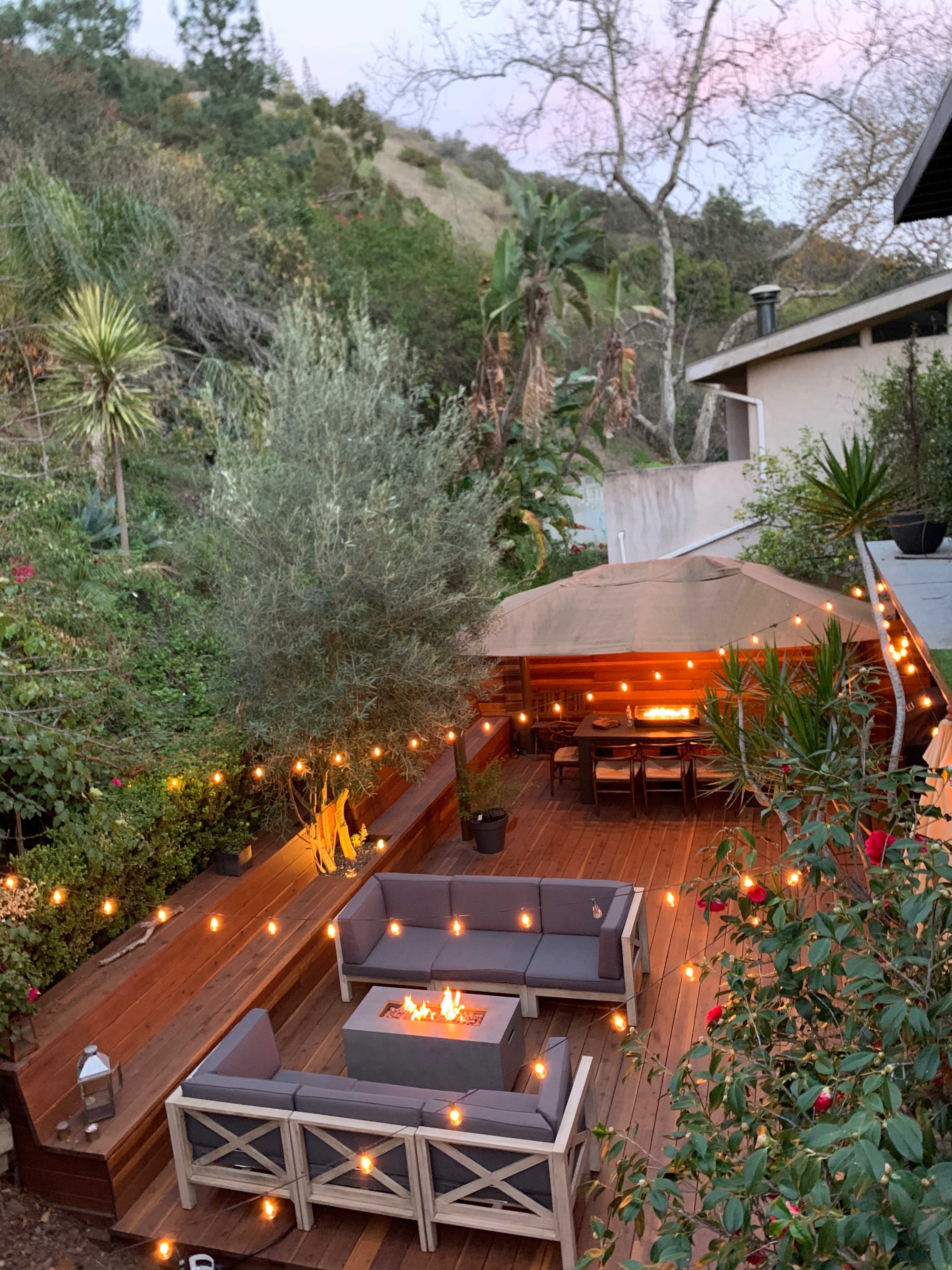 A wooden patio features a fire pit surrounded by seating, with string lights illuminating the area amidst greenery and a tented seating space.