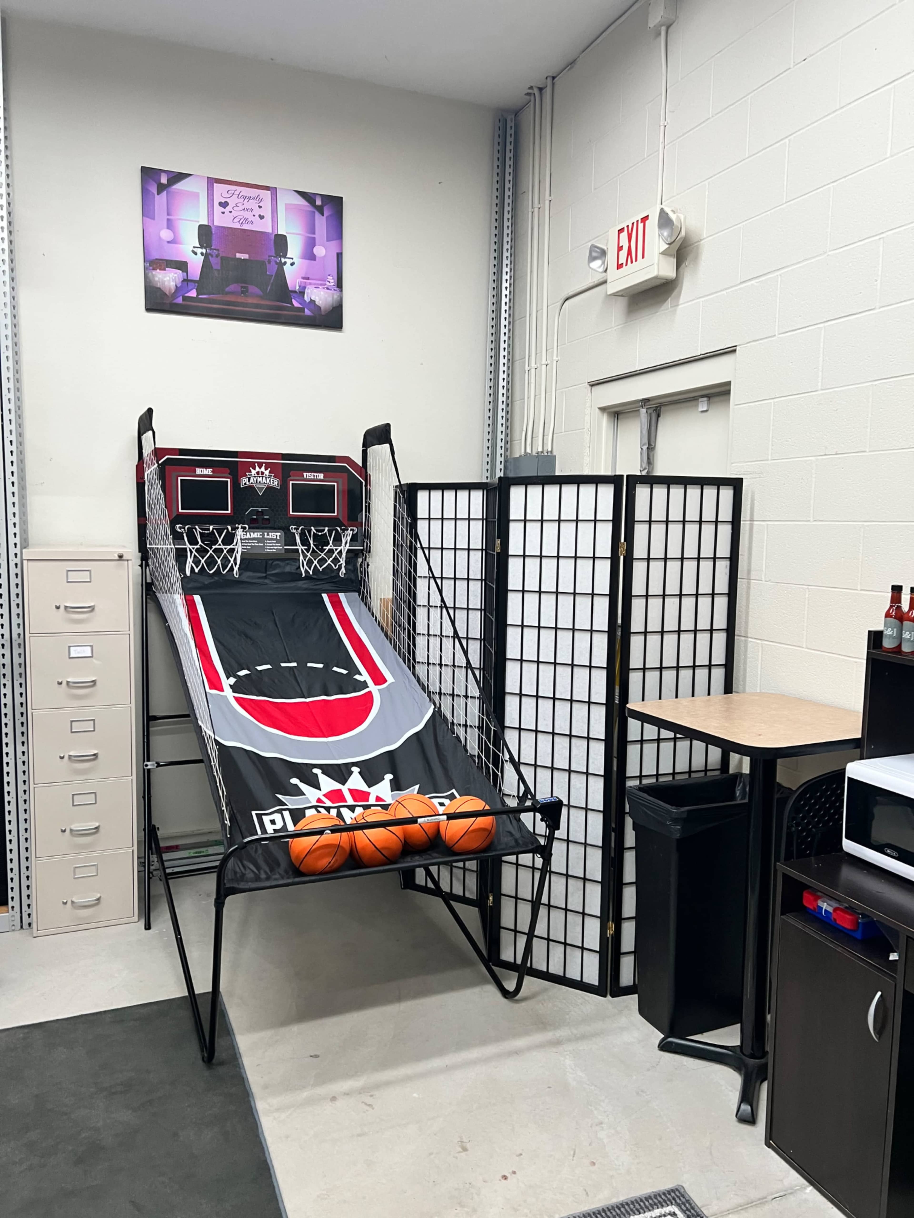 An indoor space featuring a basketball shooting arcade game against a wall, with a folding screen nearby and storage furniture in the background.