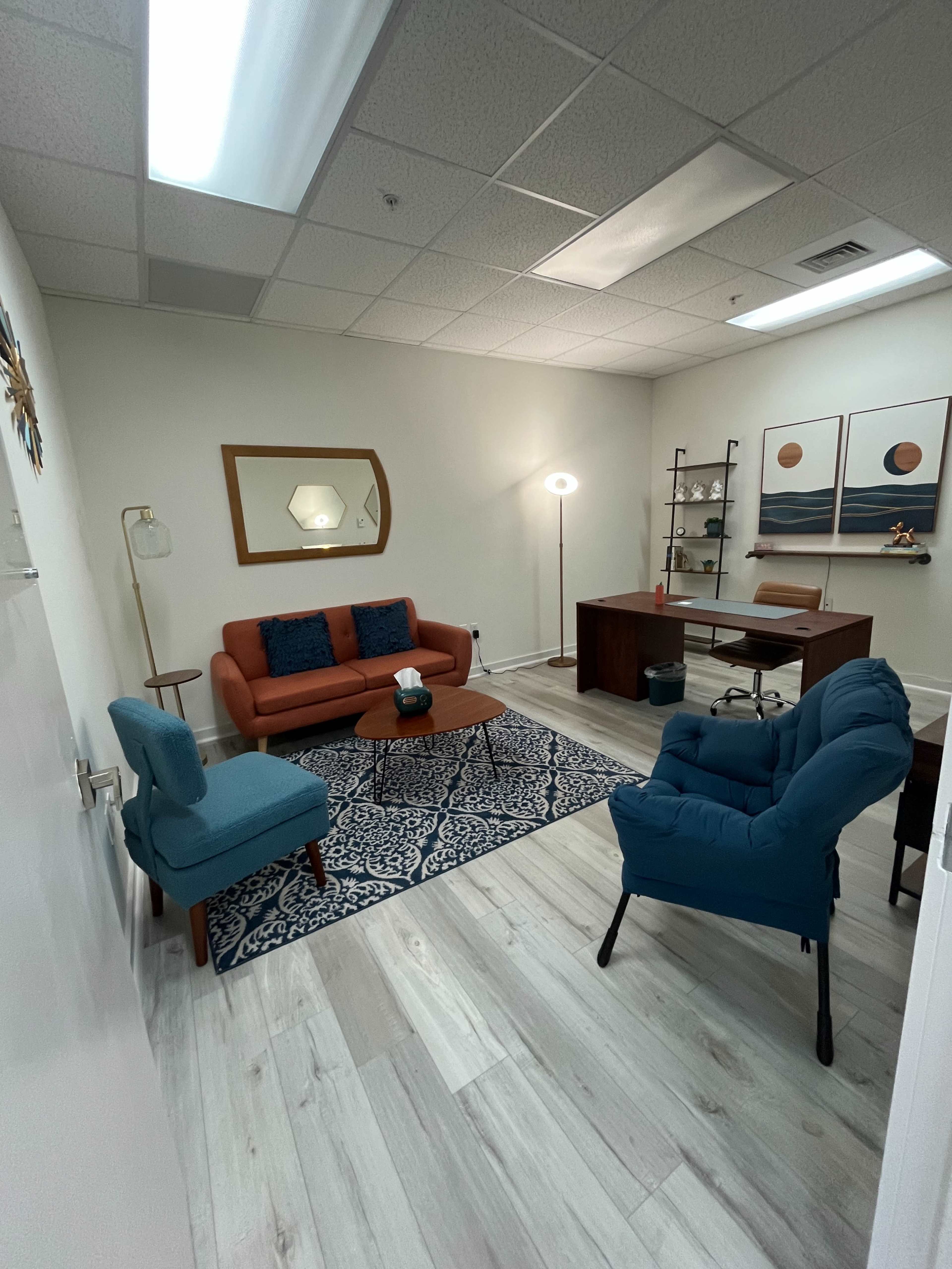 A modern office space featuring a brown desk, a blue rug, a seating area with an orange couch and two blue chairs, and wall art above the desk.