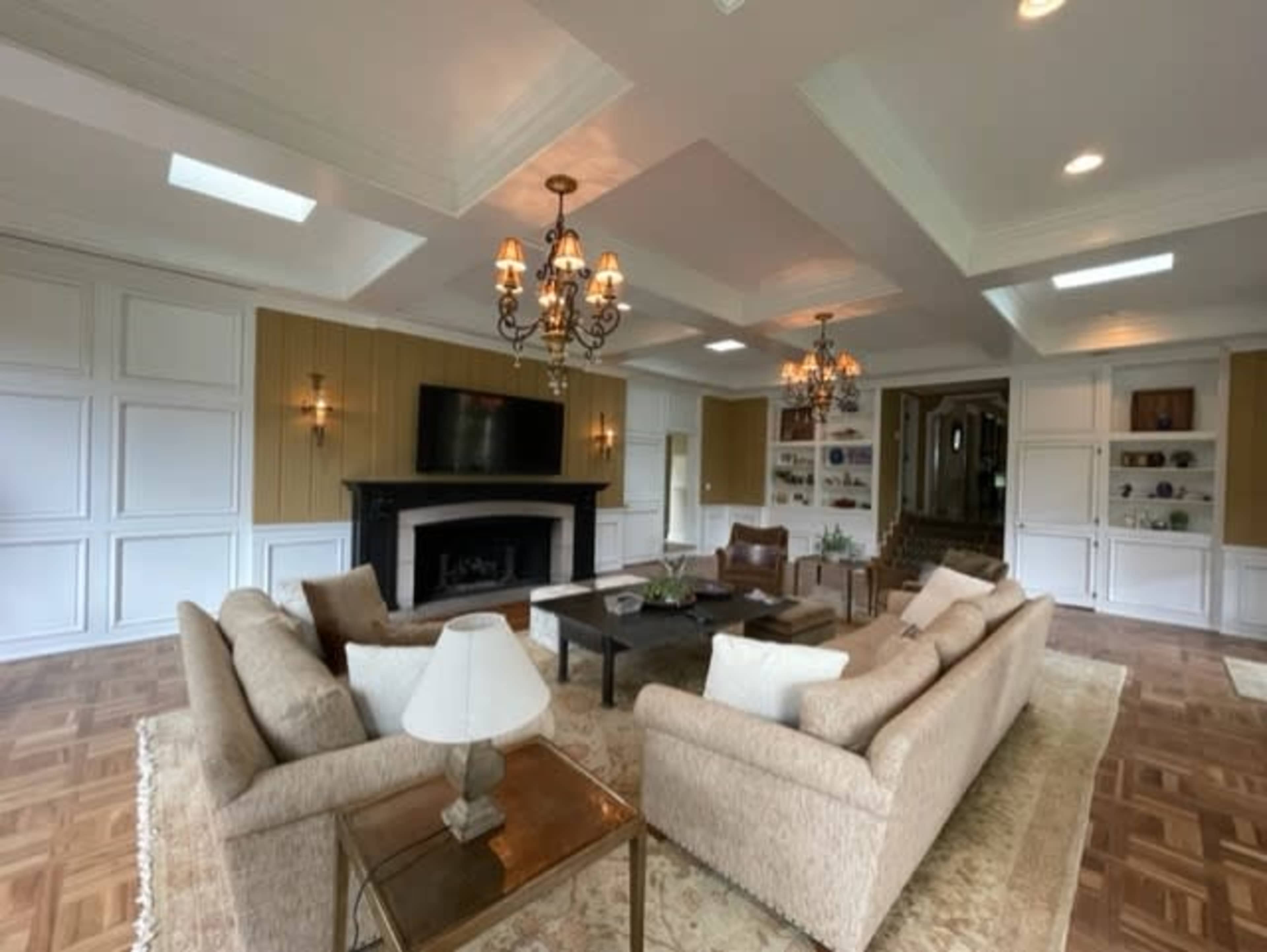 A spacious living room with two chandeliers, a large television mounted on a wall above a fireplace, and a seating arrangement centered around a coffee table.