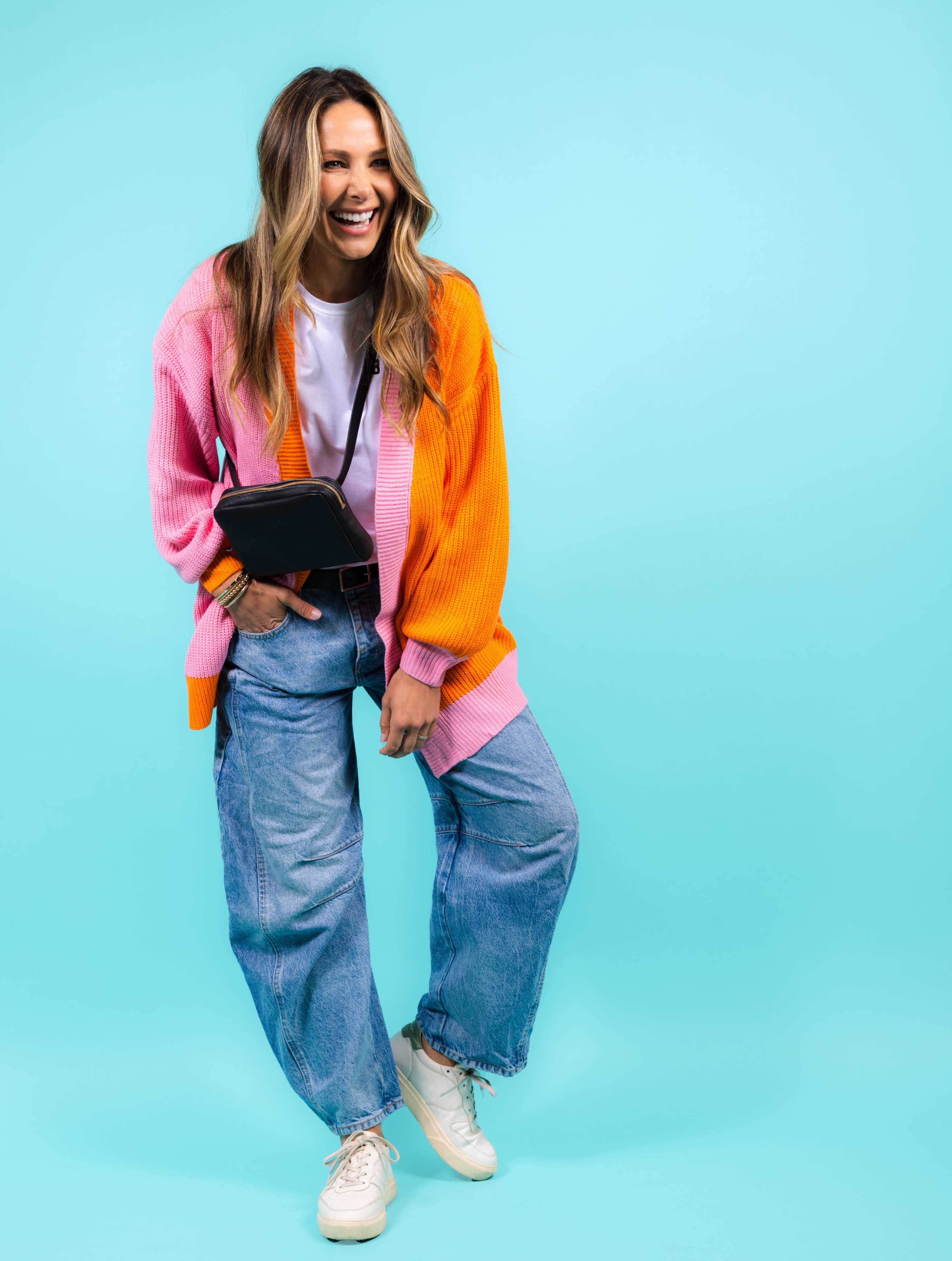 A woman wearing a colorful cardigan, white t-shirt, and baggy jeans smiles while posing against a solid teal background.