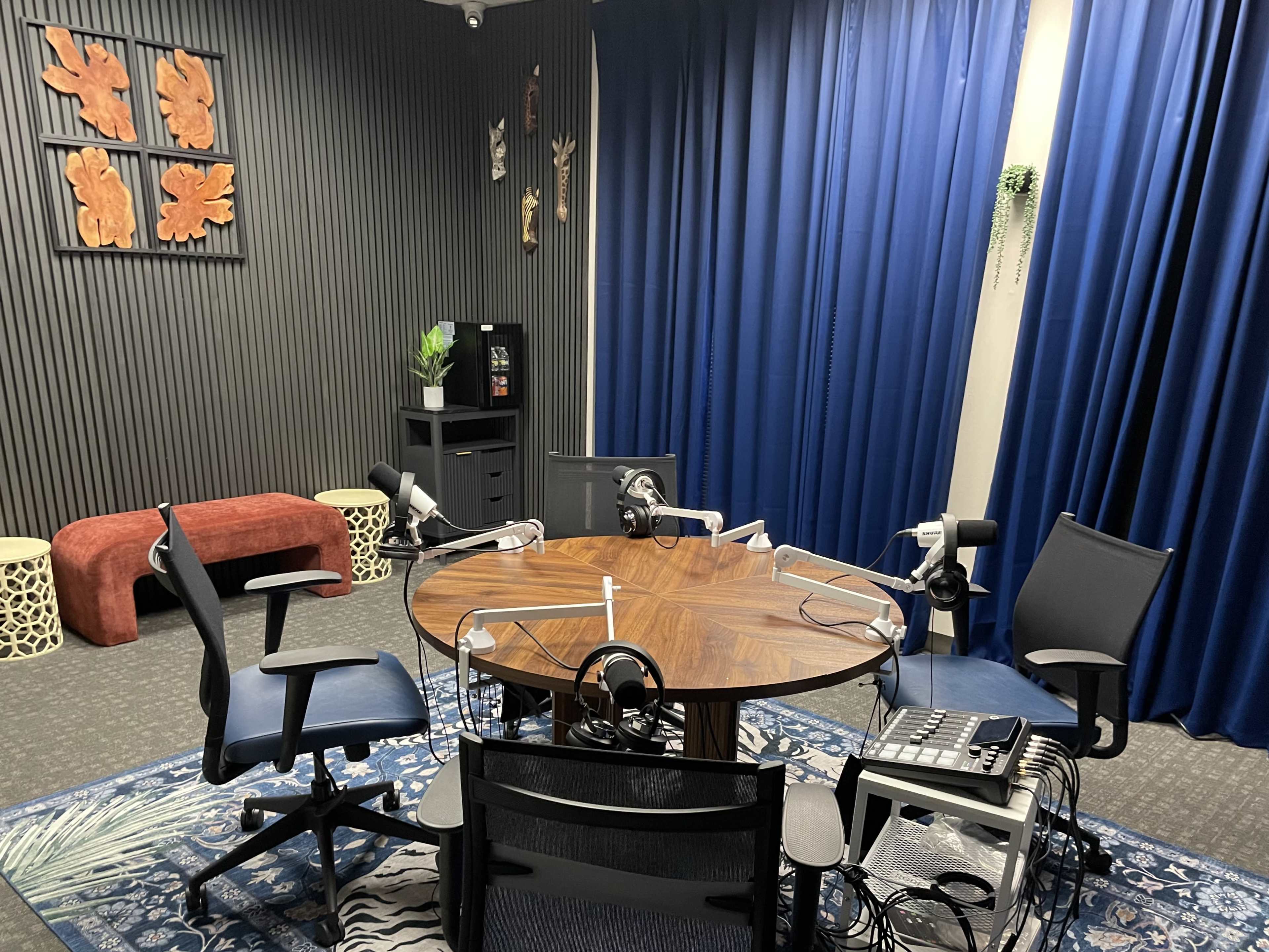 A small podcast studio features a circular table with four microphones, surrounded by chairs, against a backdrop of blue curtains and decorative wall art.