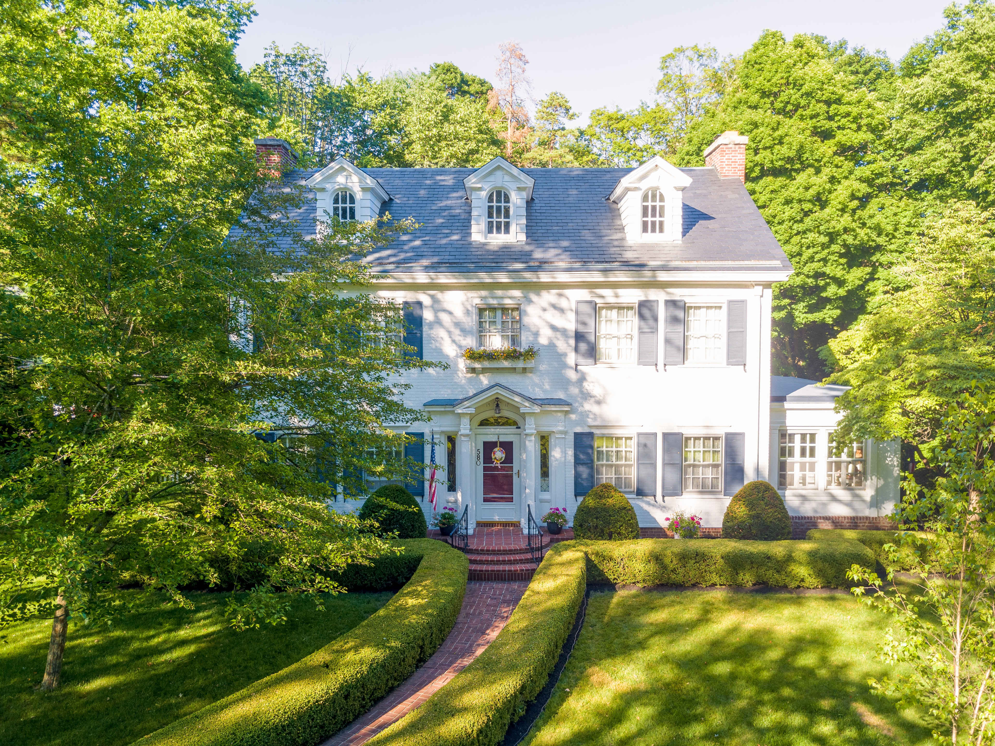 A large white house with shutters and a central entrance is surrounded by well-manicured lawns and greenery.