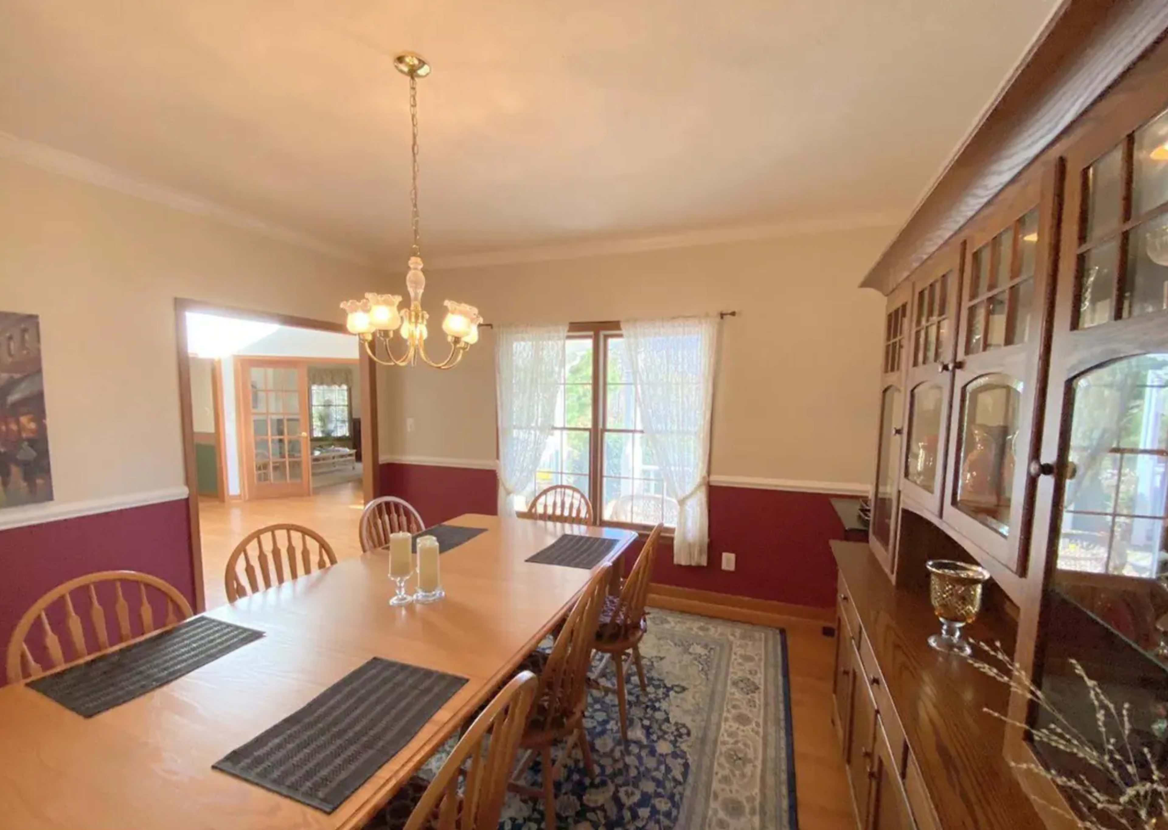 A dining room features a wooden table with eight chairs, a chandelier above, and a sideboard against the wall near large windows.