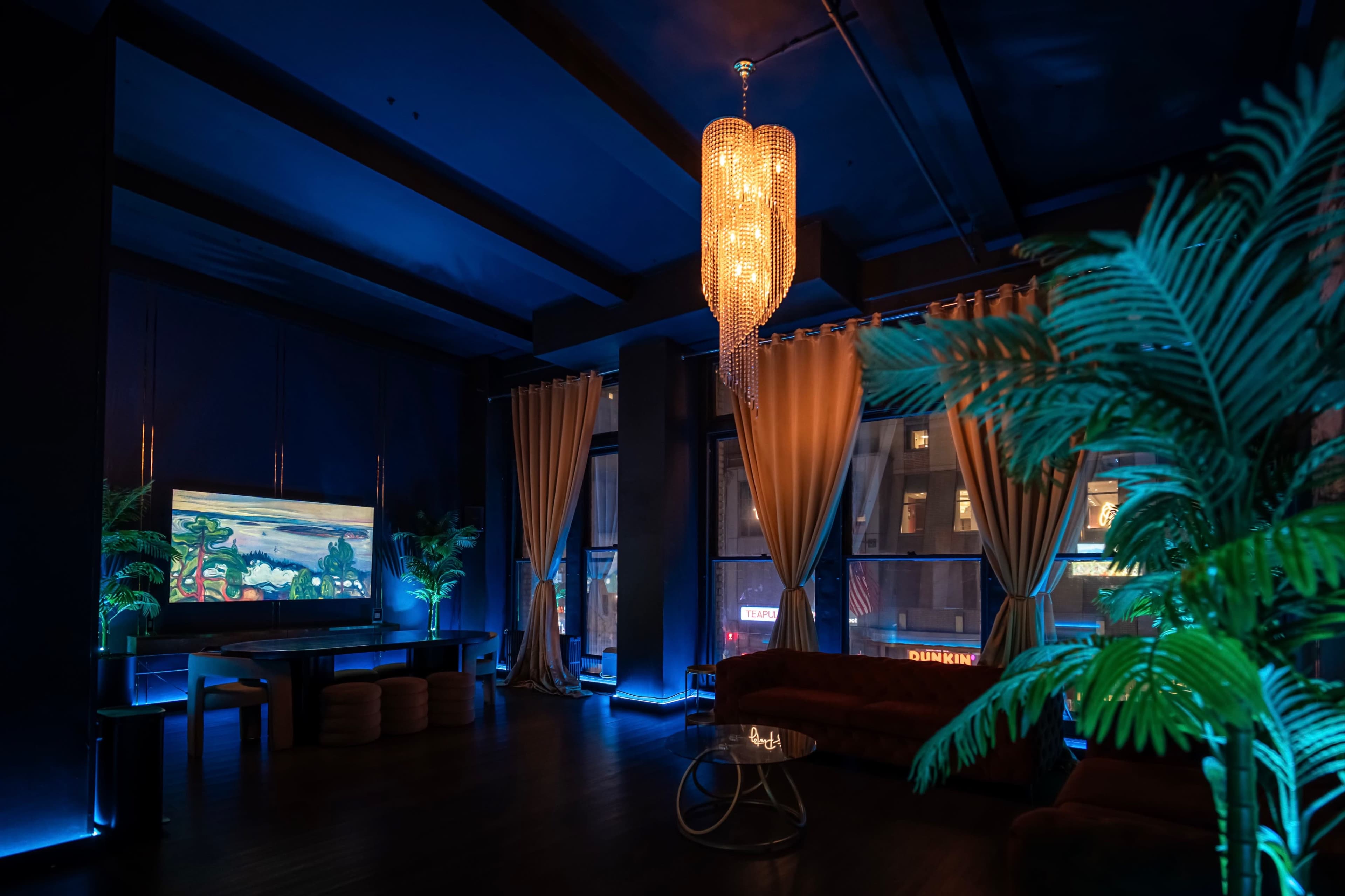 The image depicts a dimly lit lounge area featuring a large painting on the wall, plush seating, and decorative plants, with a prominent chandelier illuminating the space.