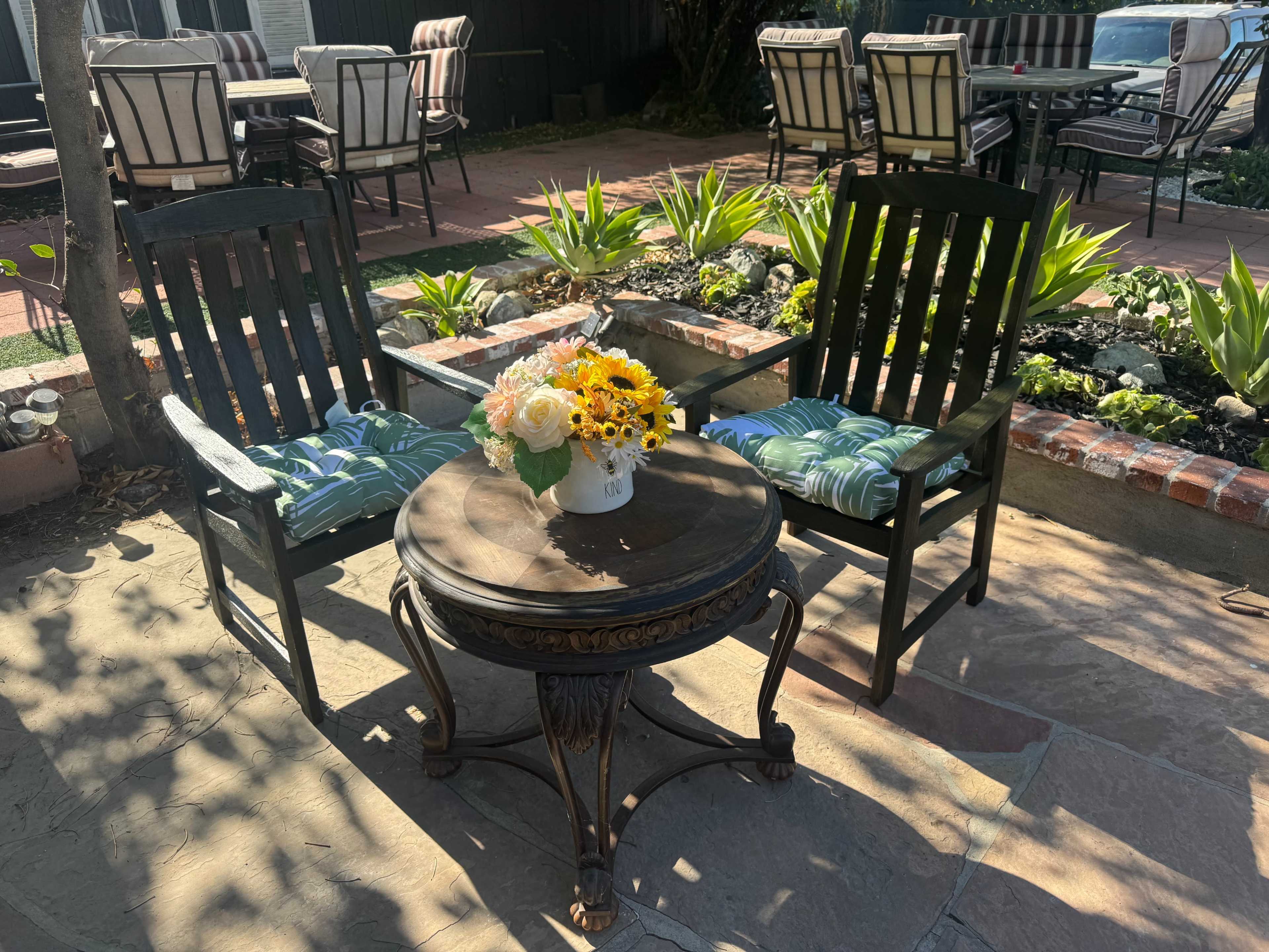 Two black wooden chairs with patterned cushions flank a round table adorned with a white vase of flowers, set in a sunlit patio surrounded by green plants.