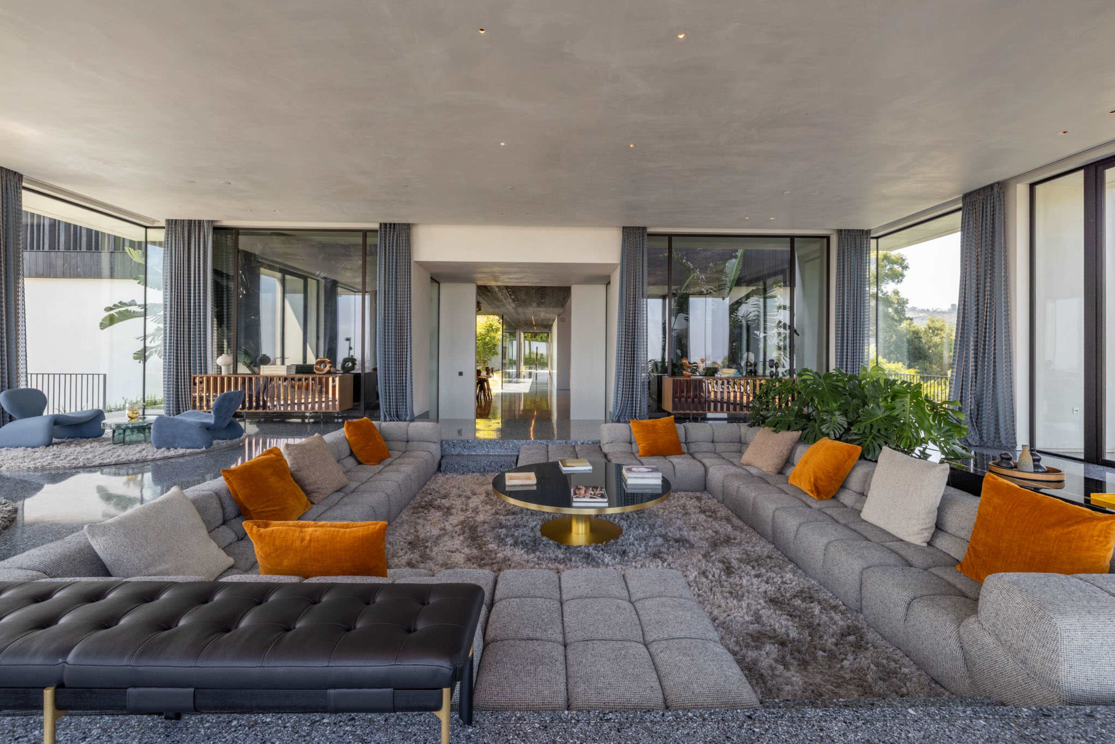 The image shows a spacious modern living room with a large sectional sofa, orange accent pillows, a low coffee table, and large windows allowing natural light.