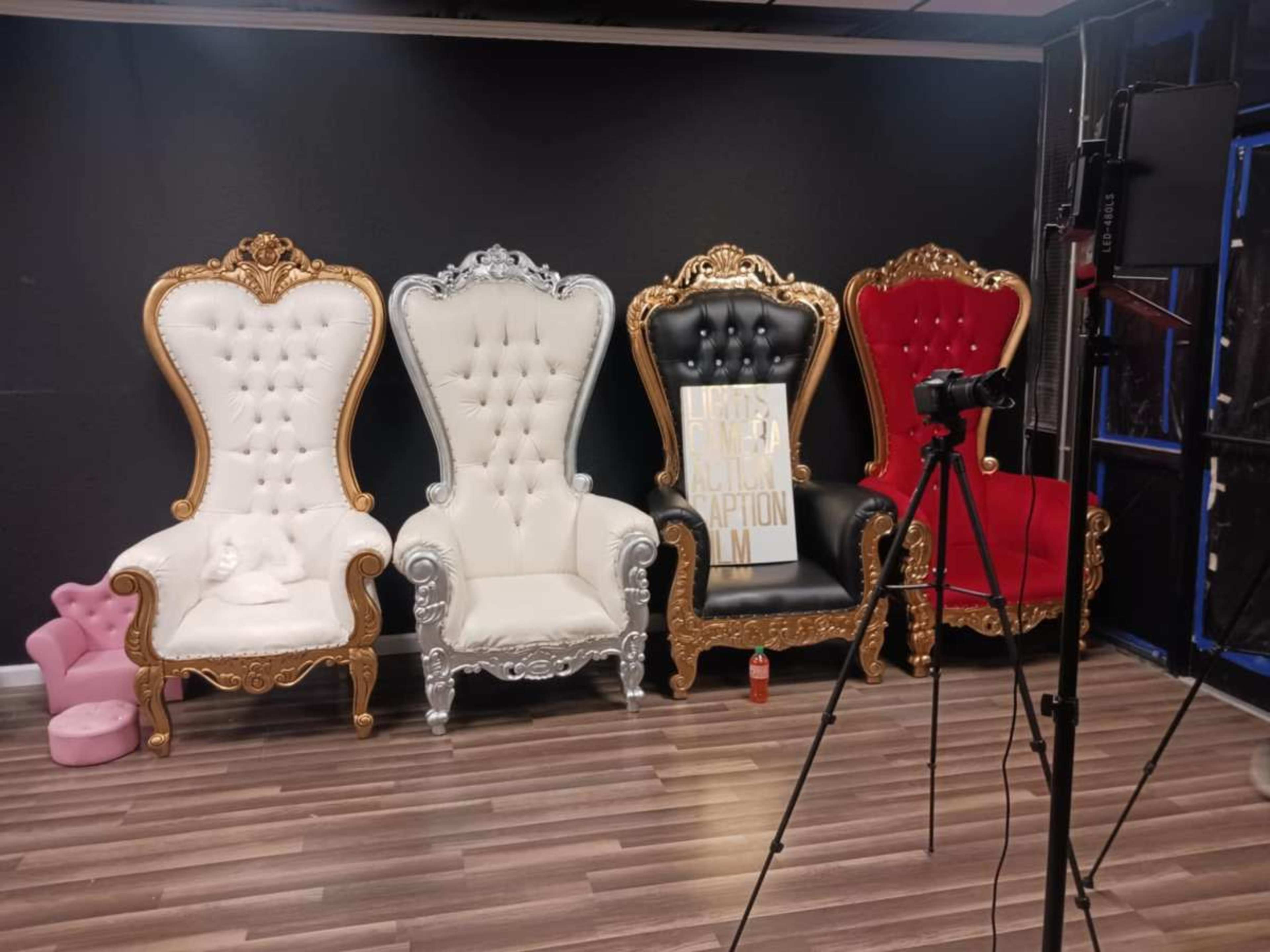 The image shows four ornate chairs in different colors—white, silver, black, and red—arranged against a dark wall, with a camera and tripod positioned in front of them.