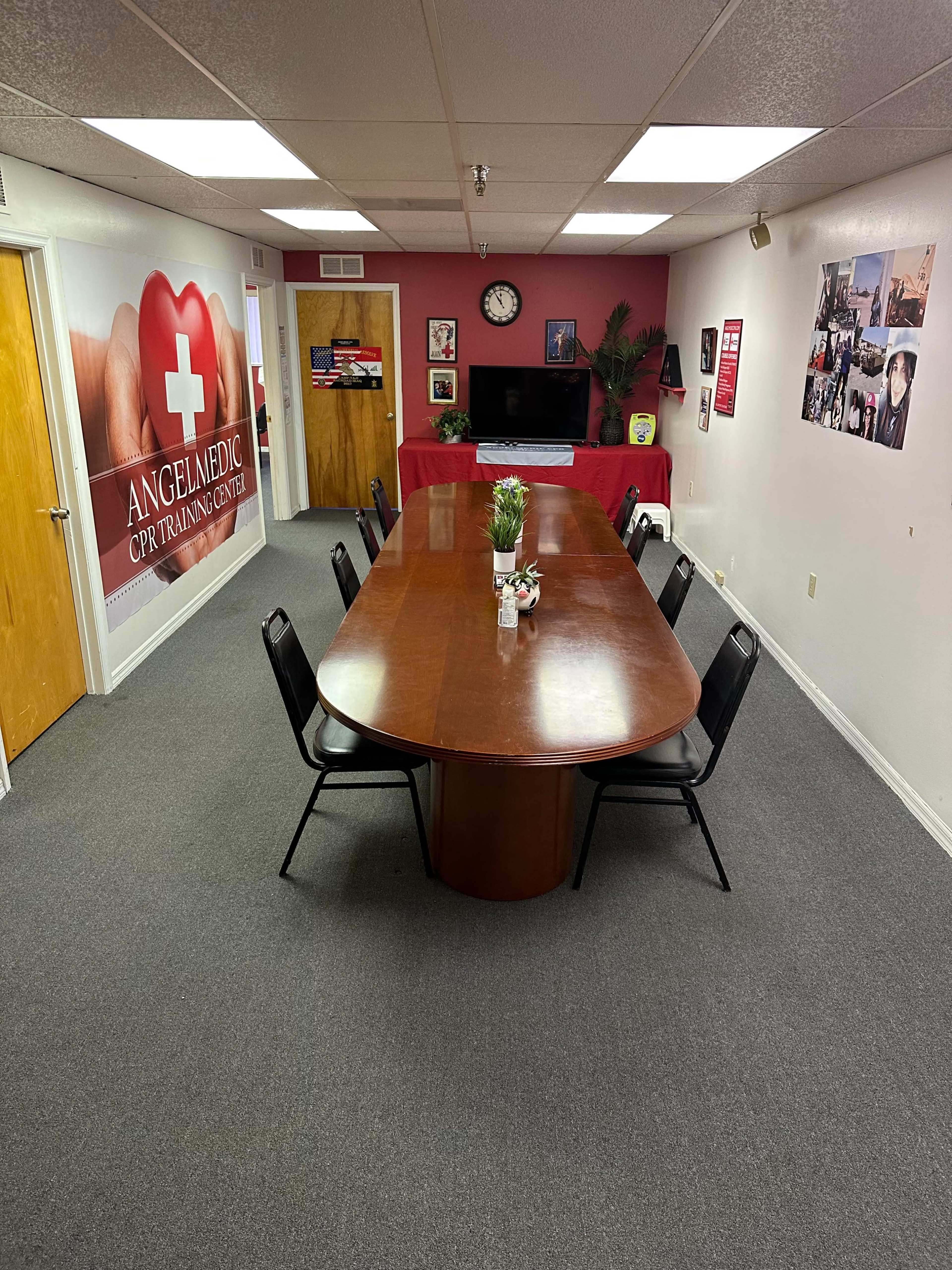 A spacious training room with a large wooden table surrounded by black chairs, decorated with a clock and various posters on the walls.
