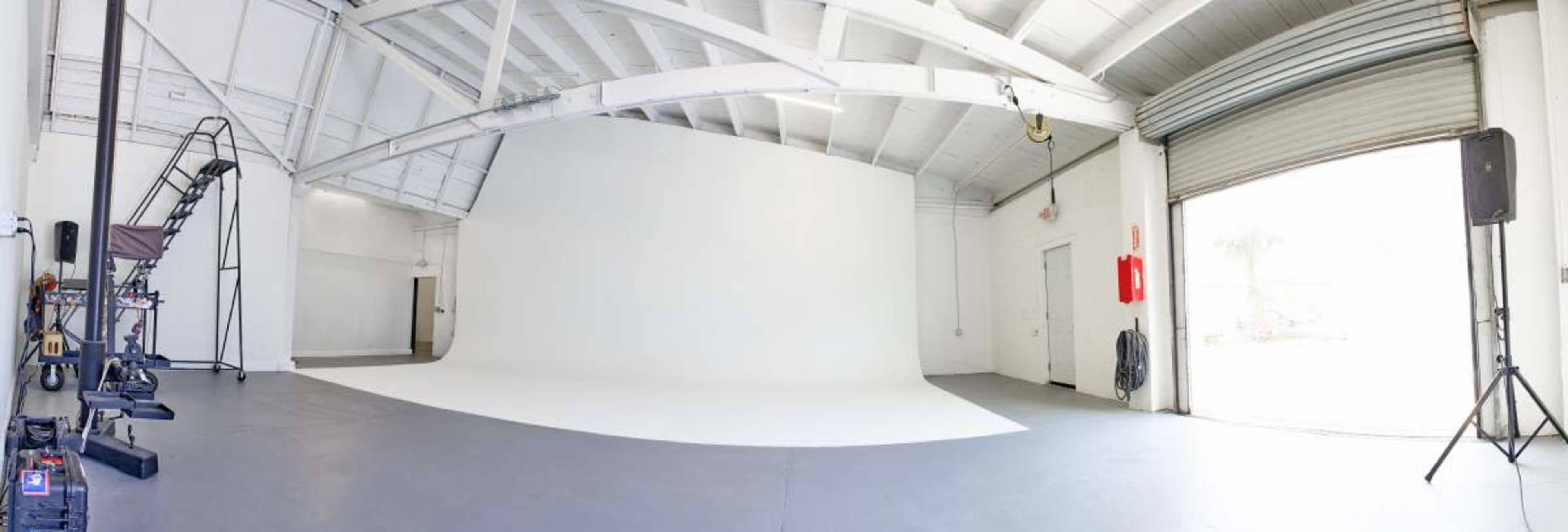 The image shows a spacious photography studio with a seamless white backdrop, overhead lighting, and a large opening for natural light.