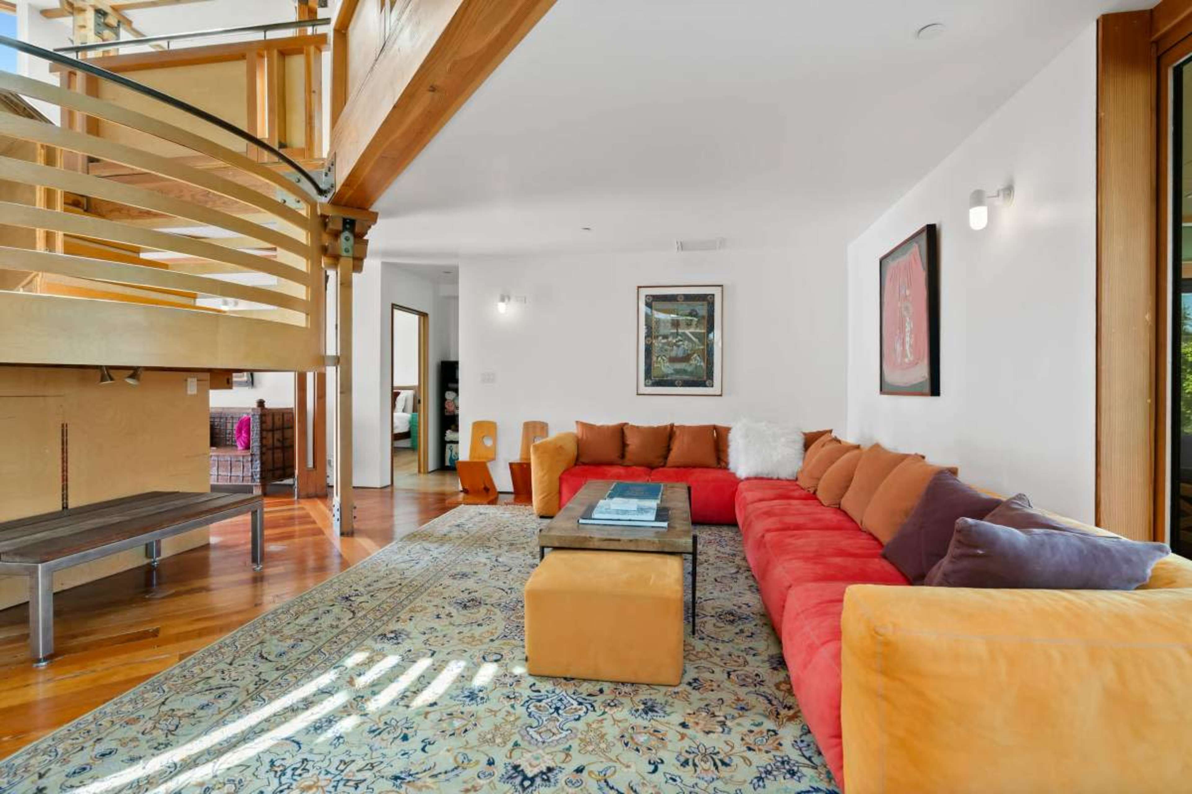 A spacious living area features a large, colorful sectional sofa, a glass coffee table, and wooden accents, with a staircase visible in the background.