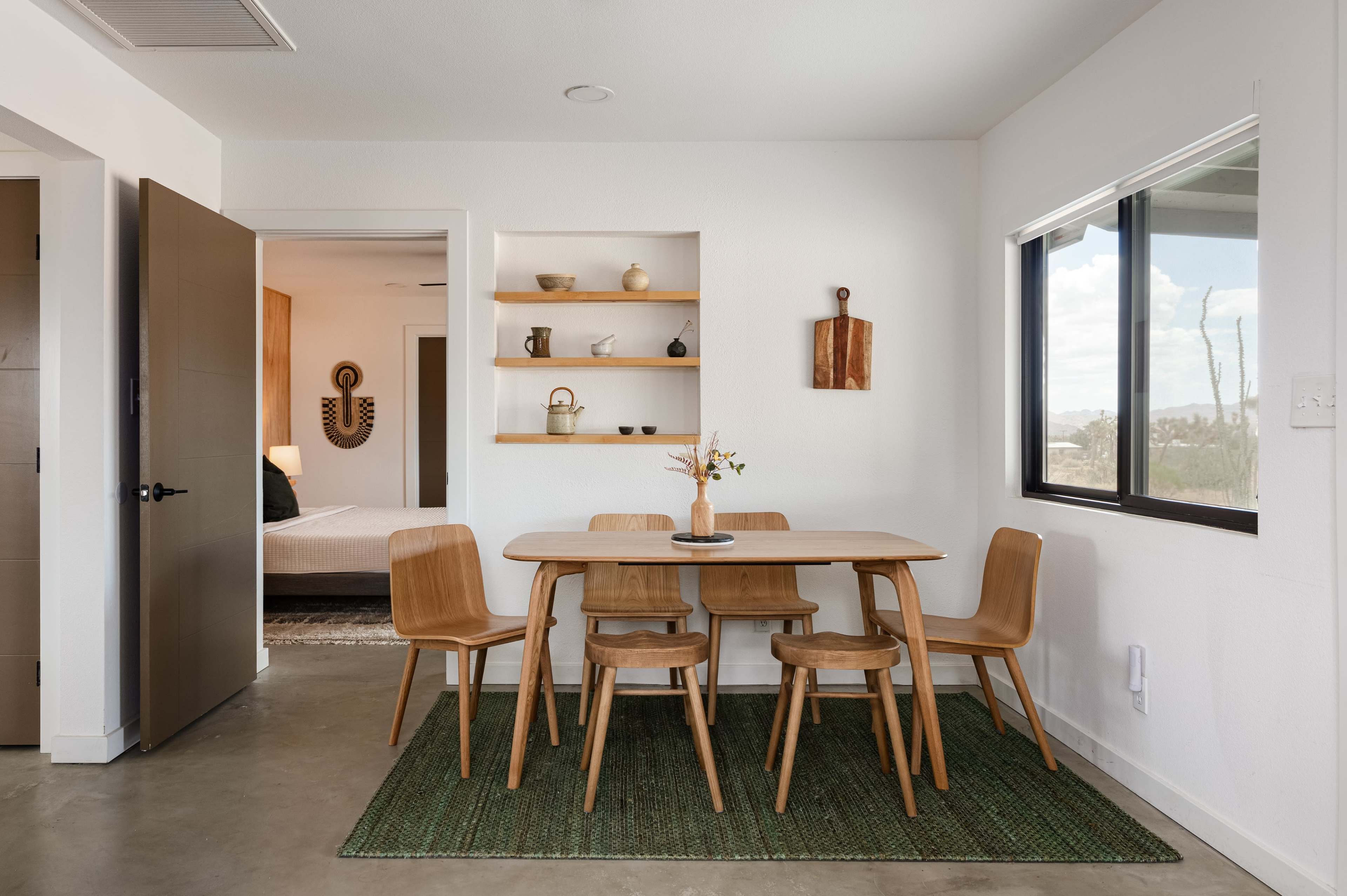 A wooden dining table with four chairs is set on a green rug in a bright room featuring a wall shelf with decorative items and a doorway leading to another space.