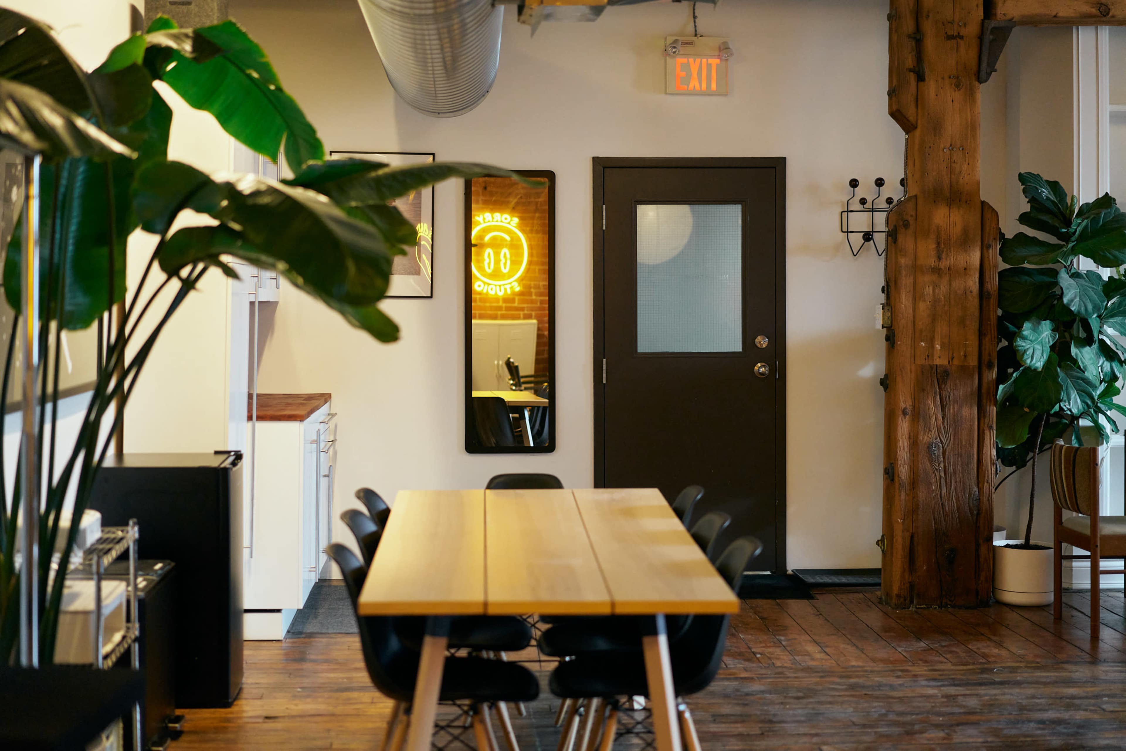 A dining area features a long wooden table, surrounded by dark chairs, with a doorway and a large mirror reflecting a neon sign in the background.