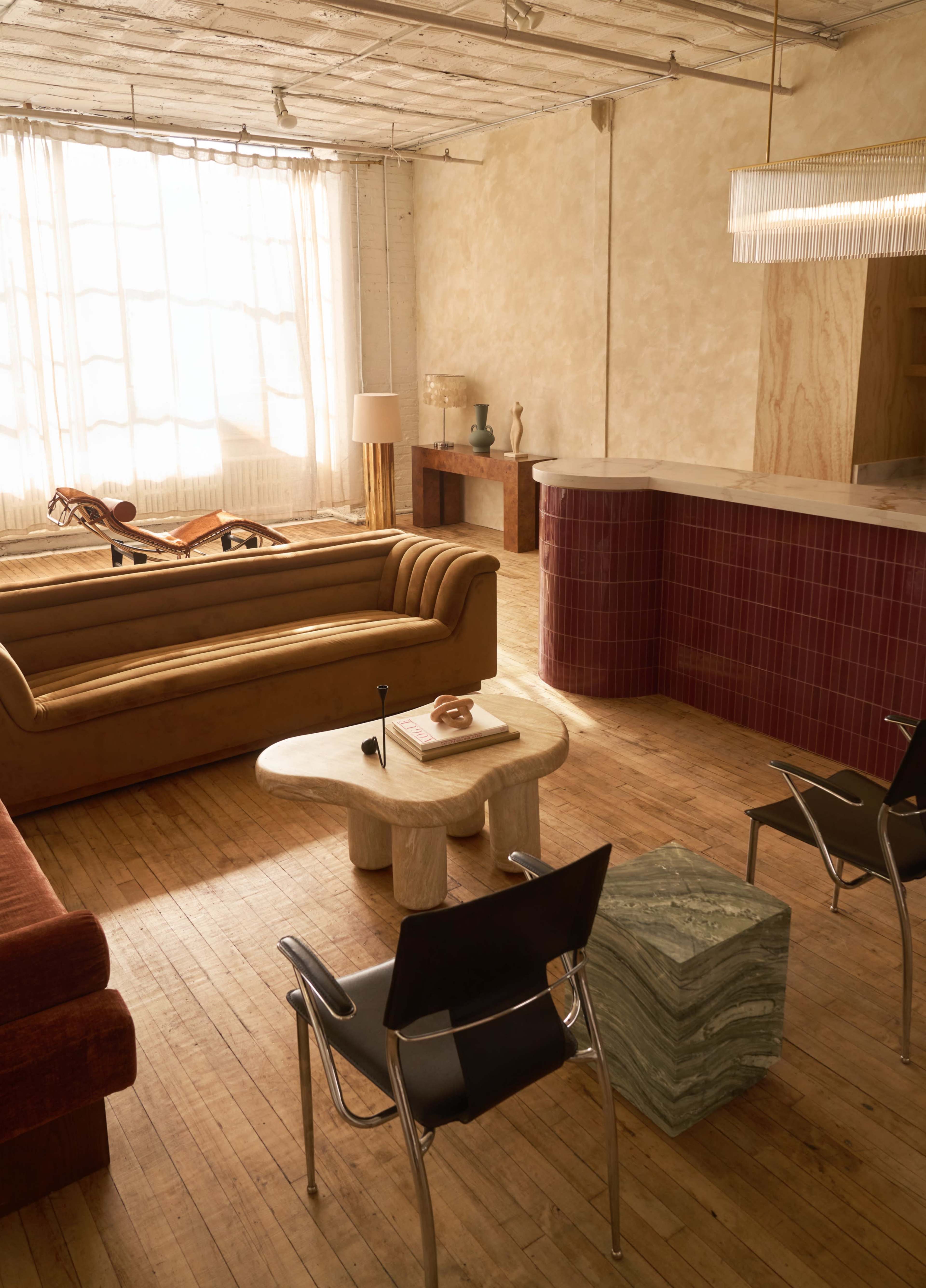 The room features a mix of vintage furniture, including a brown sofa, a marble-topped coffee table, and black chairs, all illuminated by natural light from large windows.
