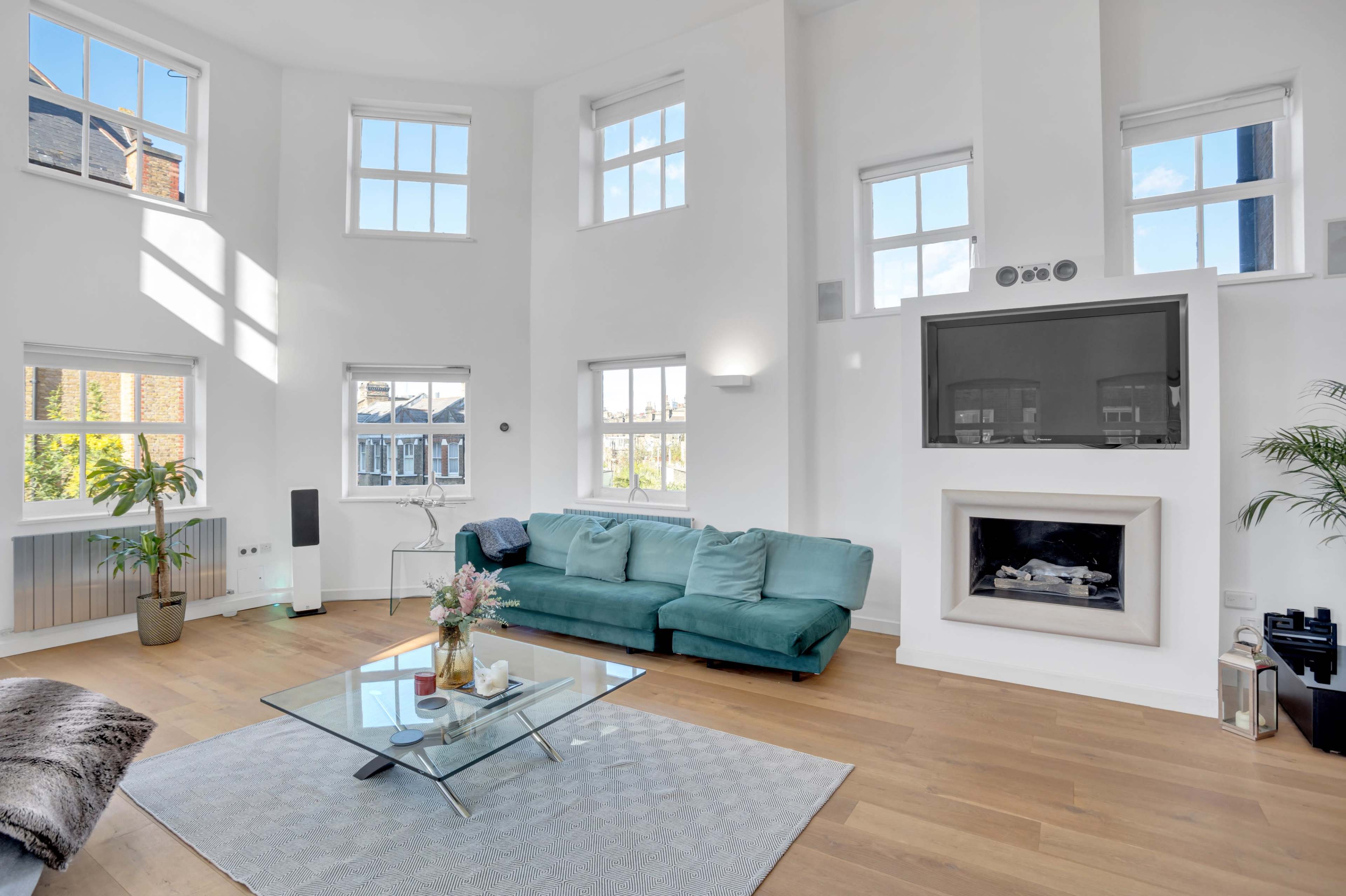 The image shows a modern living room with large windows, a green sofa, a glass coffee table, and a fireplace.