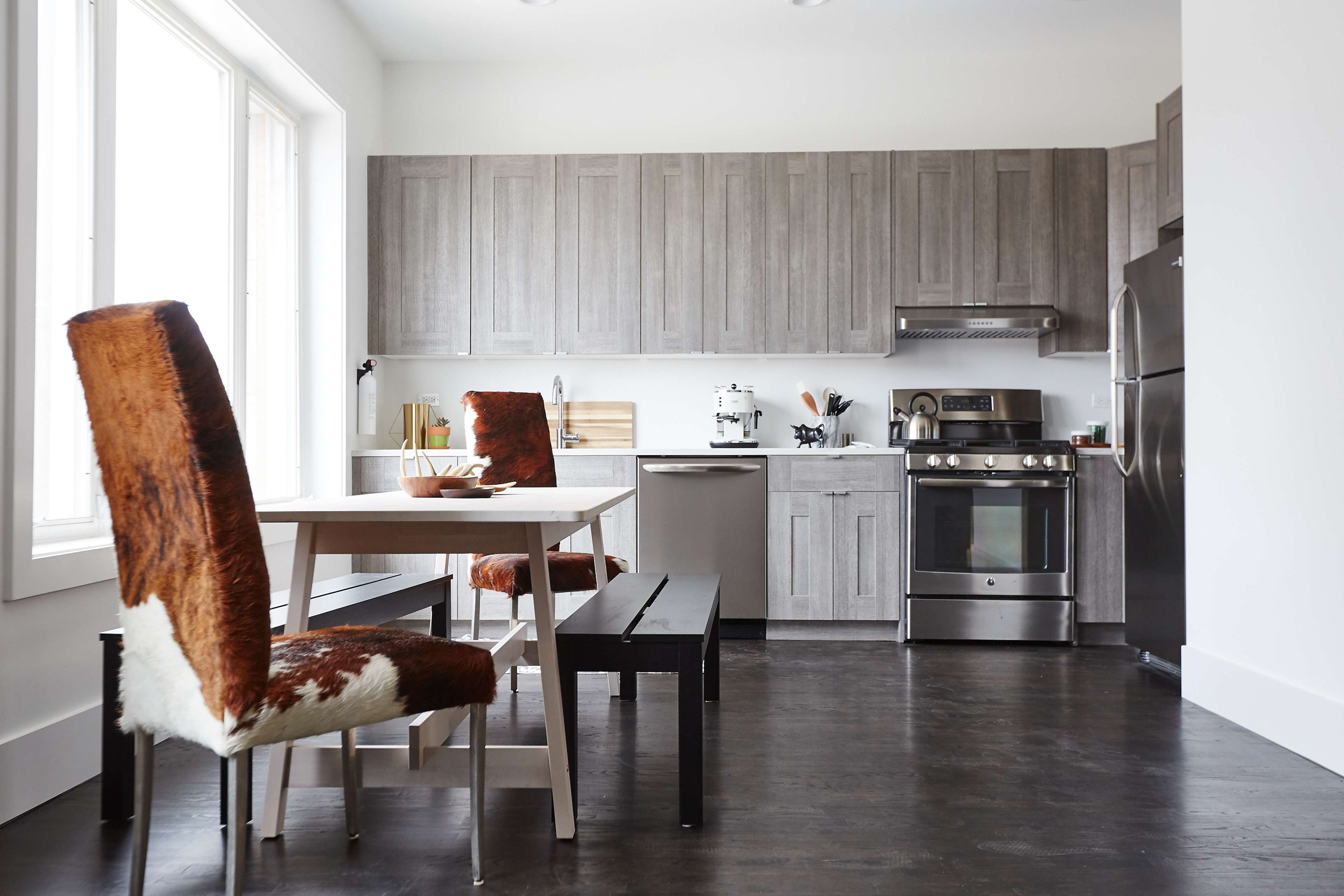A modern kitchen features gray cabinetry, stainless steel appliances, and a dining area with a table and cowhide-covered chairs.