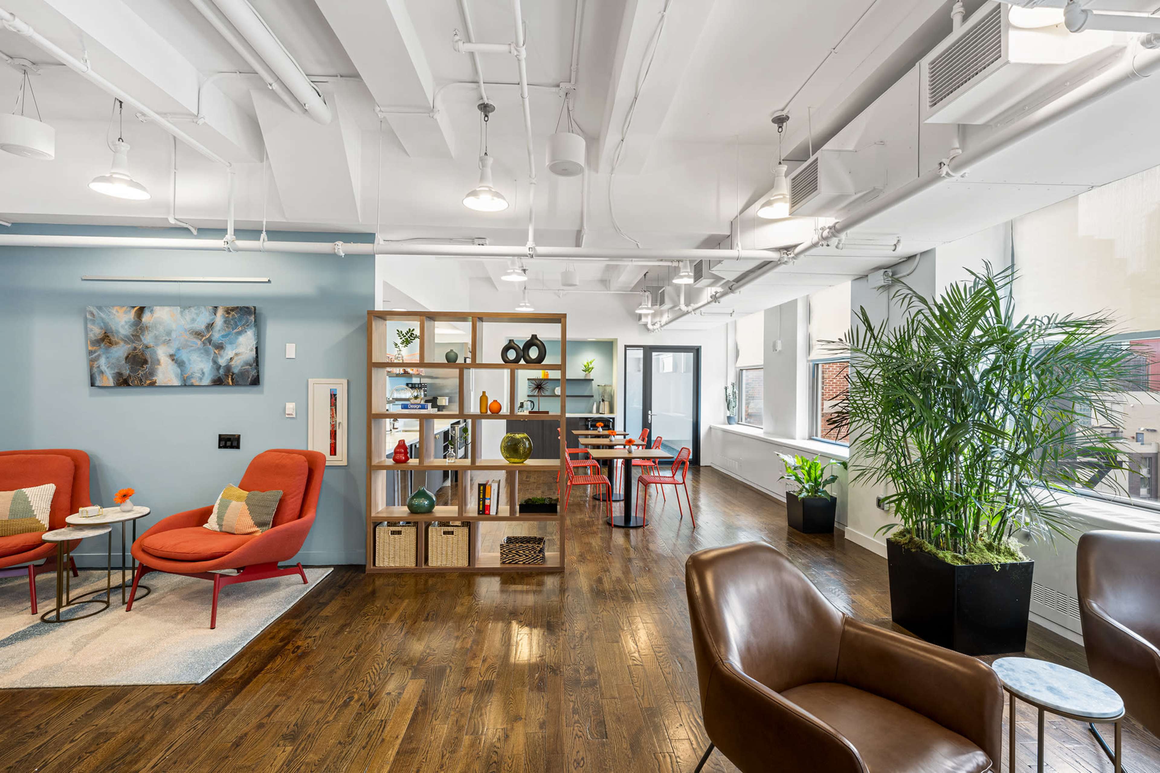 The image shows a modern office space with red upholstered chairs, a wooden bookshelf, and a plant near large windows, along with additional seating and a dining table area.