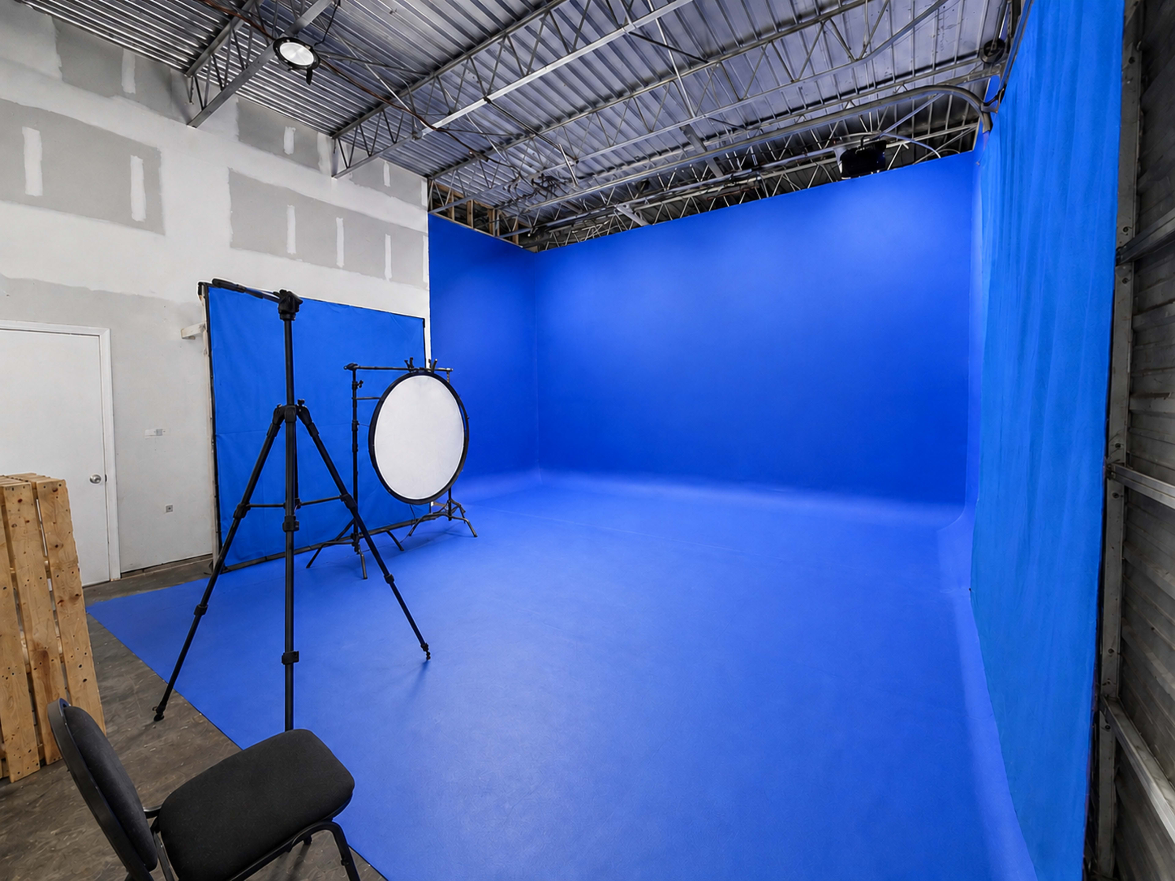 The image shows a photography studio with a solid blue backdrop, a lighting setup on a tripod, and an empty chair in the corner.