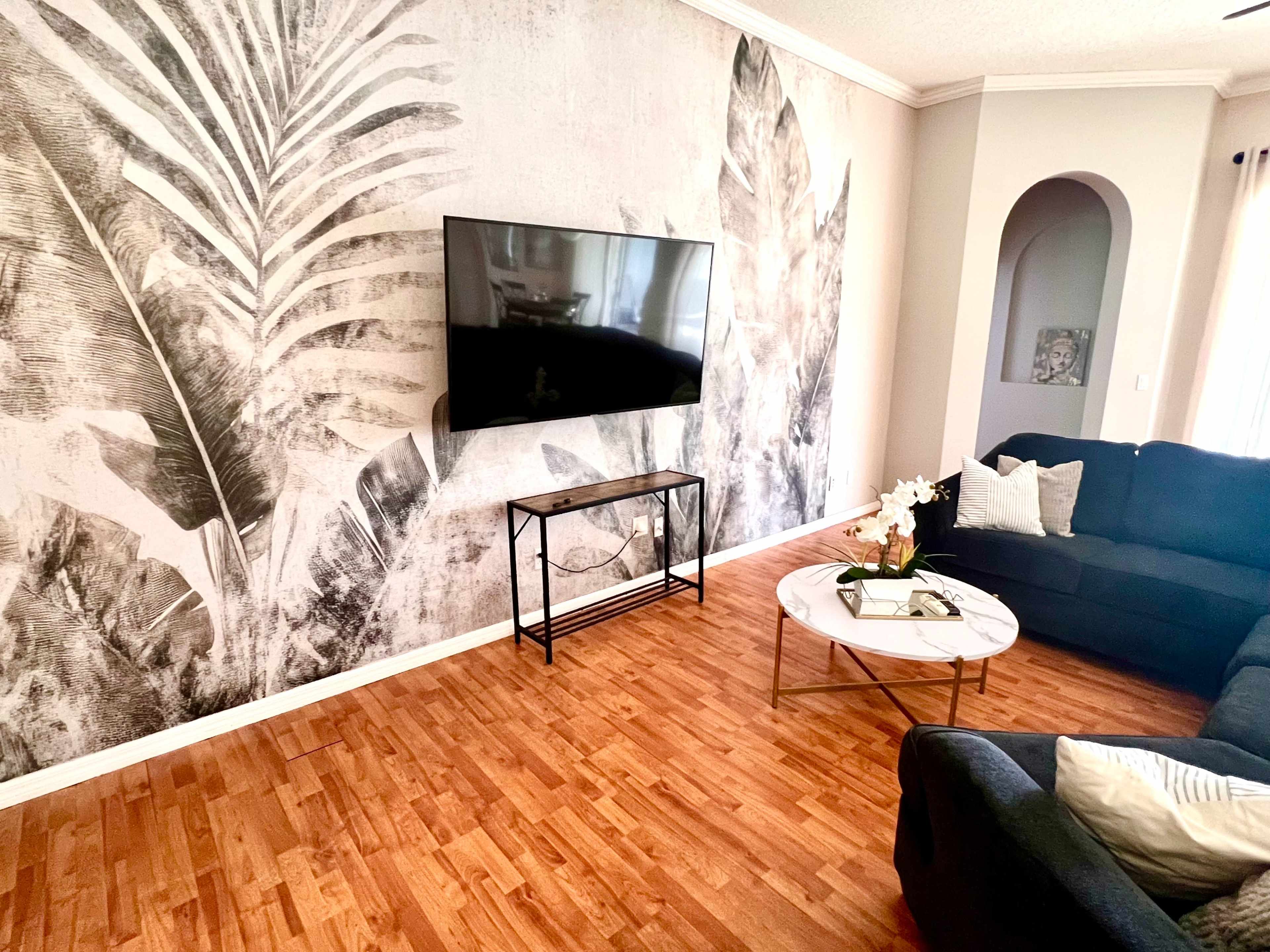 The image shows a living room with a large wall mural of tropical leaves, a mounted flat-screen TV, a coffee table, and a dark sofa.