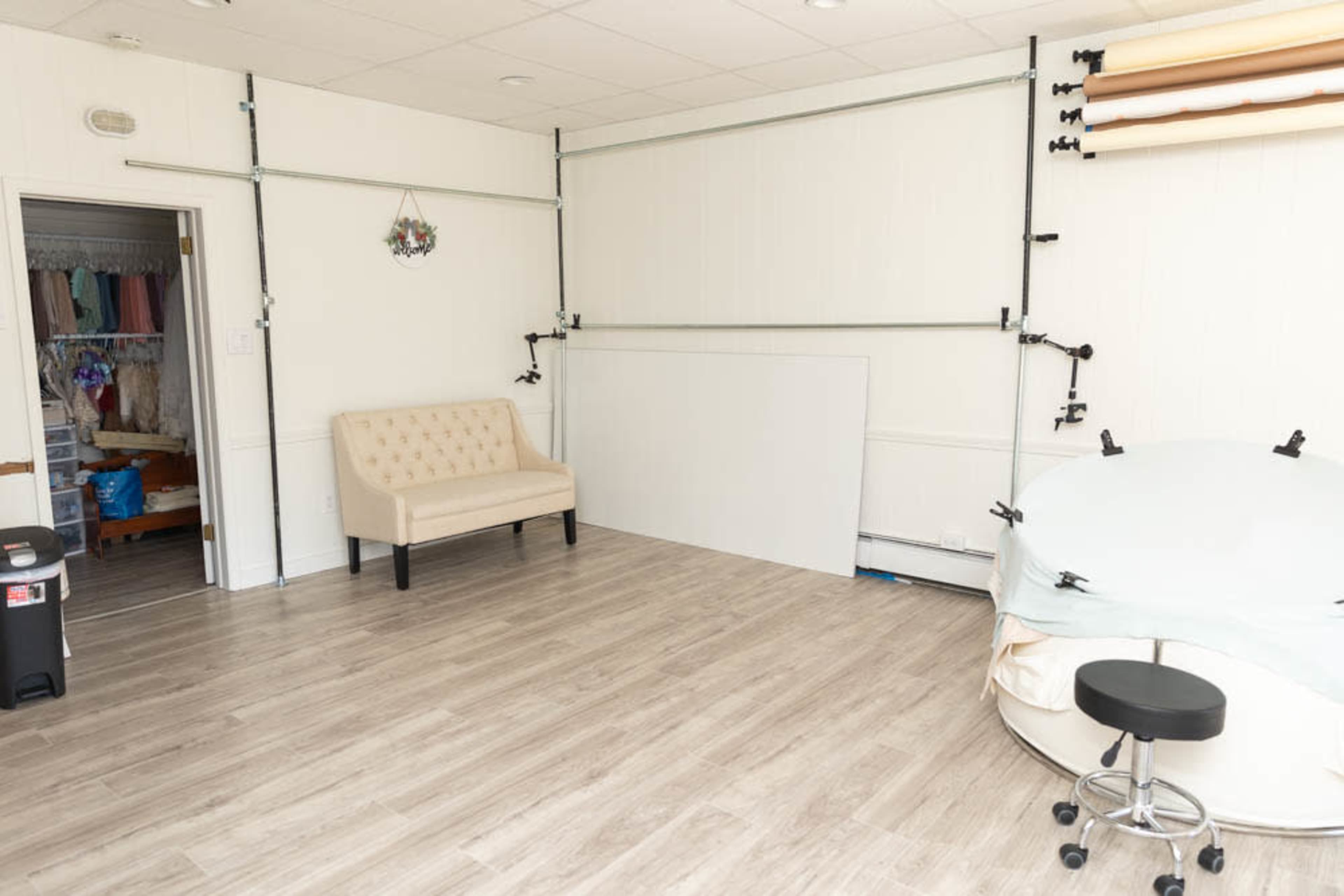 A spacious room featuring a light-colored wall, a beige couch, a circular table with a white cover, and a storage area in the background with hanging clothes.