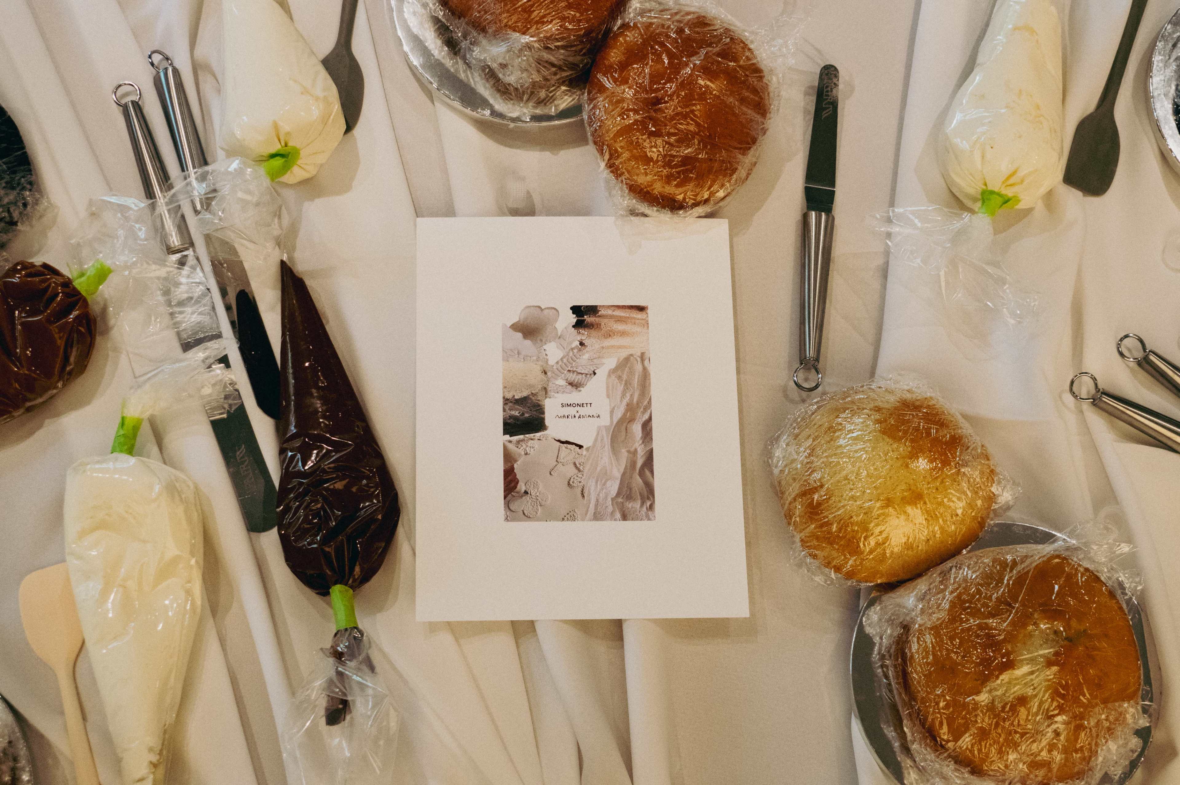 A table is spread with various frosting tools and containers, alongside wrapped buns and a central menu or guide sheet.