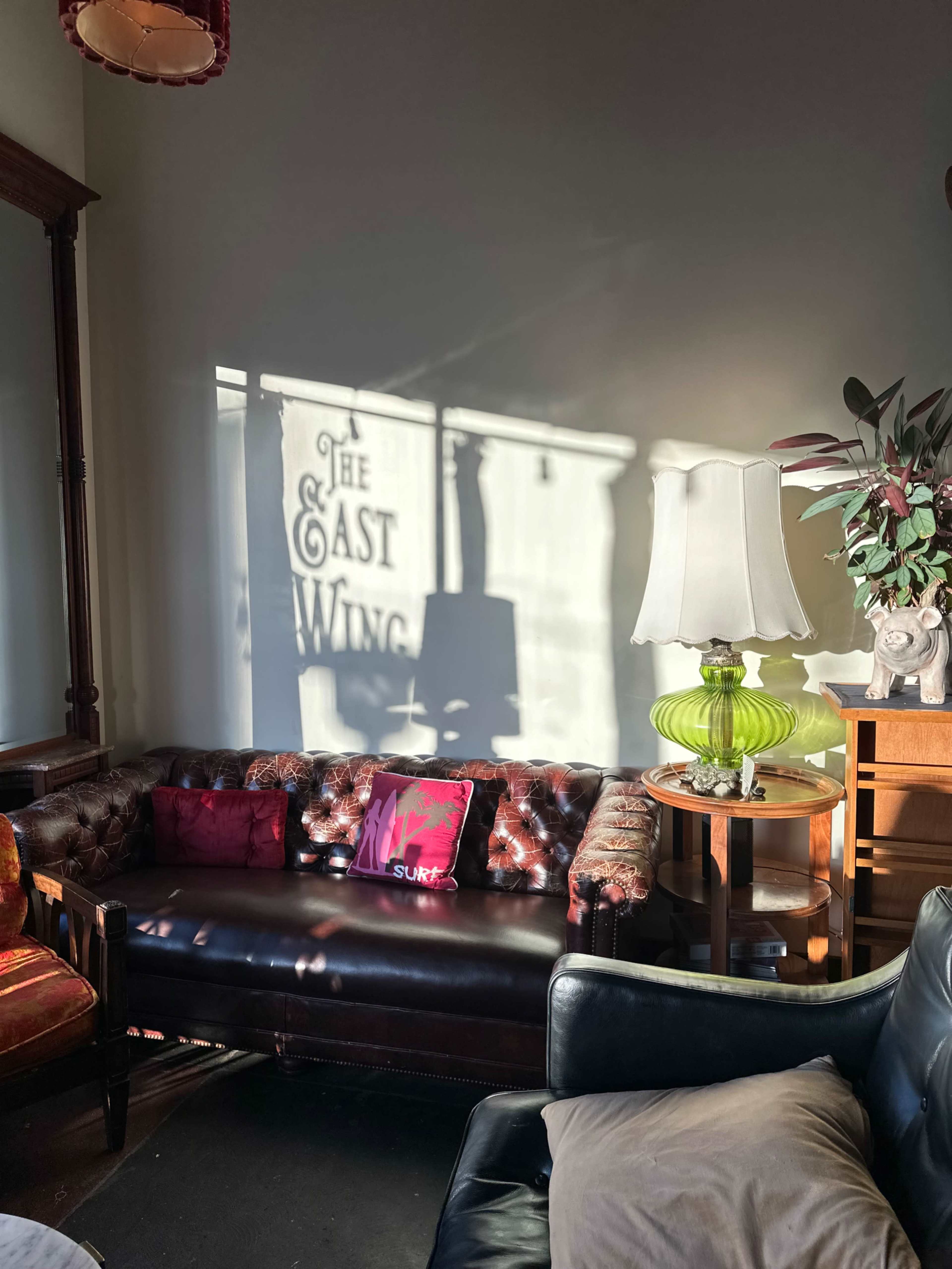 A well-furnished living space features a leather couch with colorful pillows, a lamp, and a decorative plant, while shadows create the text "THE EAST WING" on the wall.