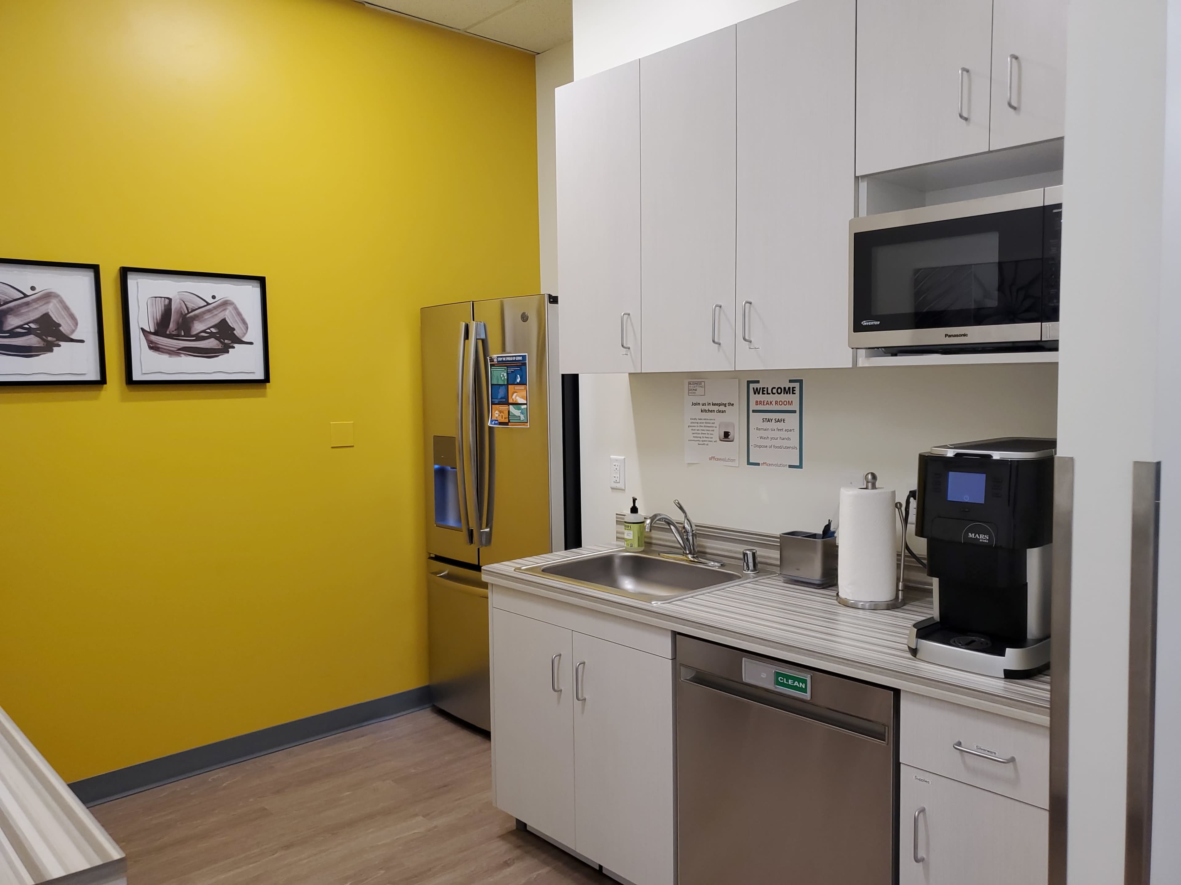 A modern kitchen equipped with a refrigerator, microwave, sink, and coffee maker against a yellow wall, with two framed pictures on display.