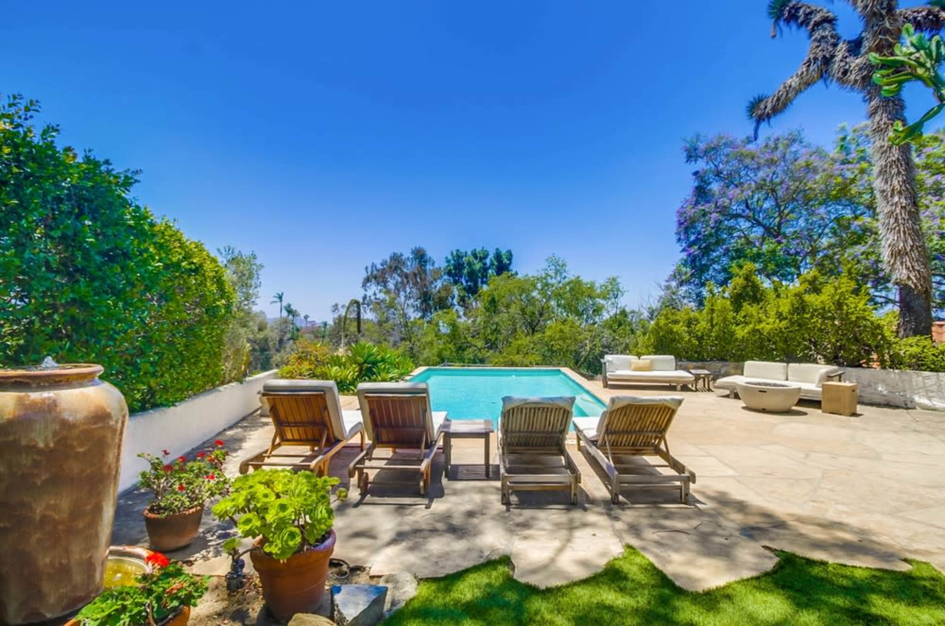The image shows a backyard with a swimming pool surrounded by lounge chairs, potted plants, and trees under a clear blue sky.