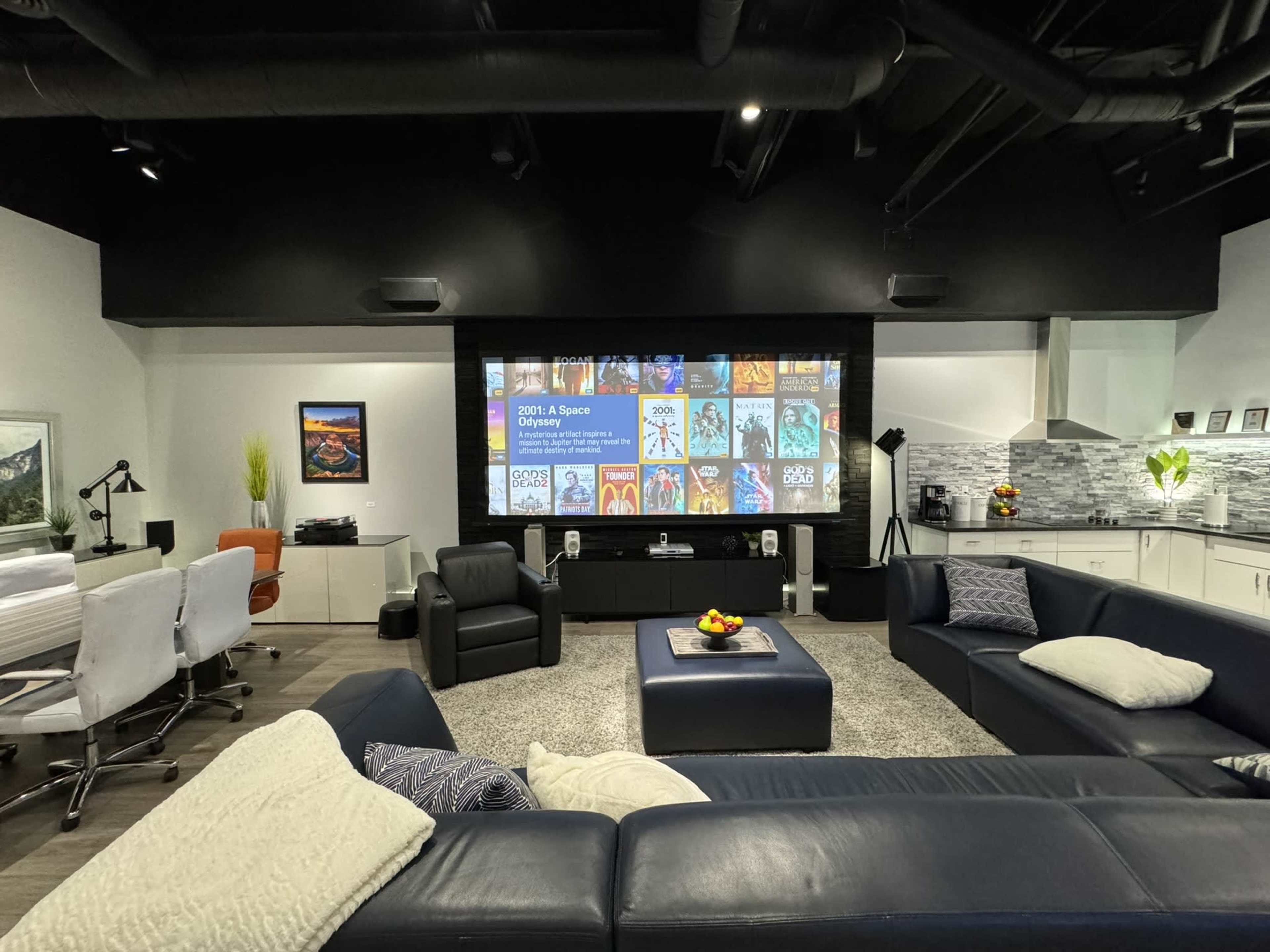 A modern home theater with a large screen displaying movie posters, surrounded by a sectional sofa, a coffee table with fruit, and a stylish workspace.