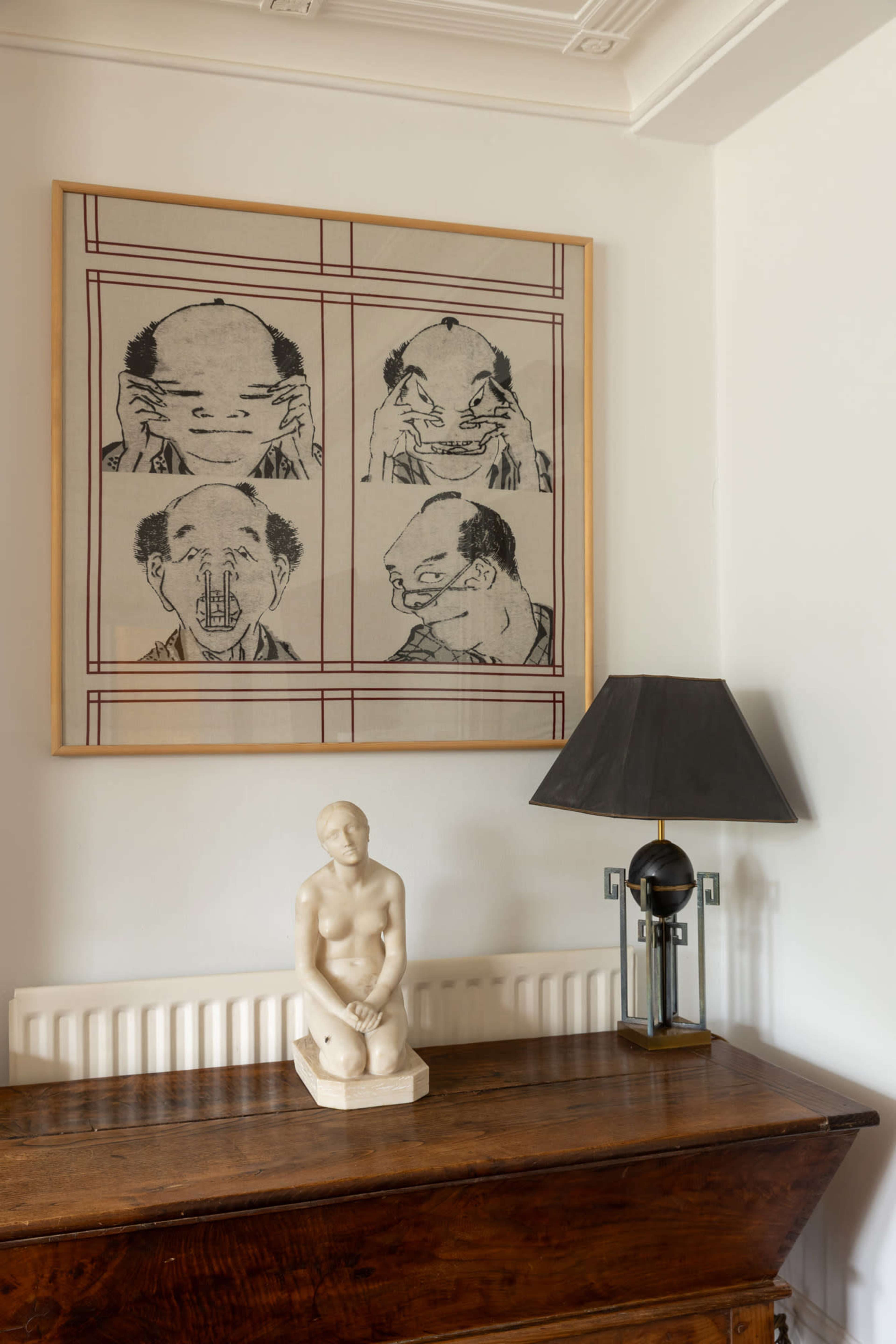 The image shows a wooden table with a sculpture of a seated figure and a lamp, underneath a large framed artwork depicting four expressive faces.