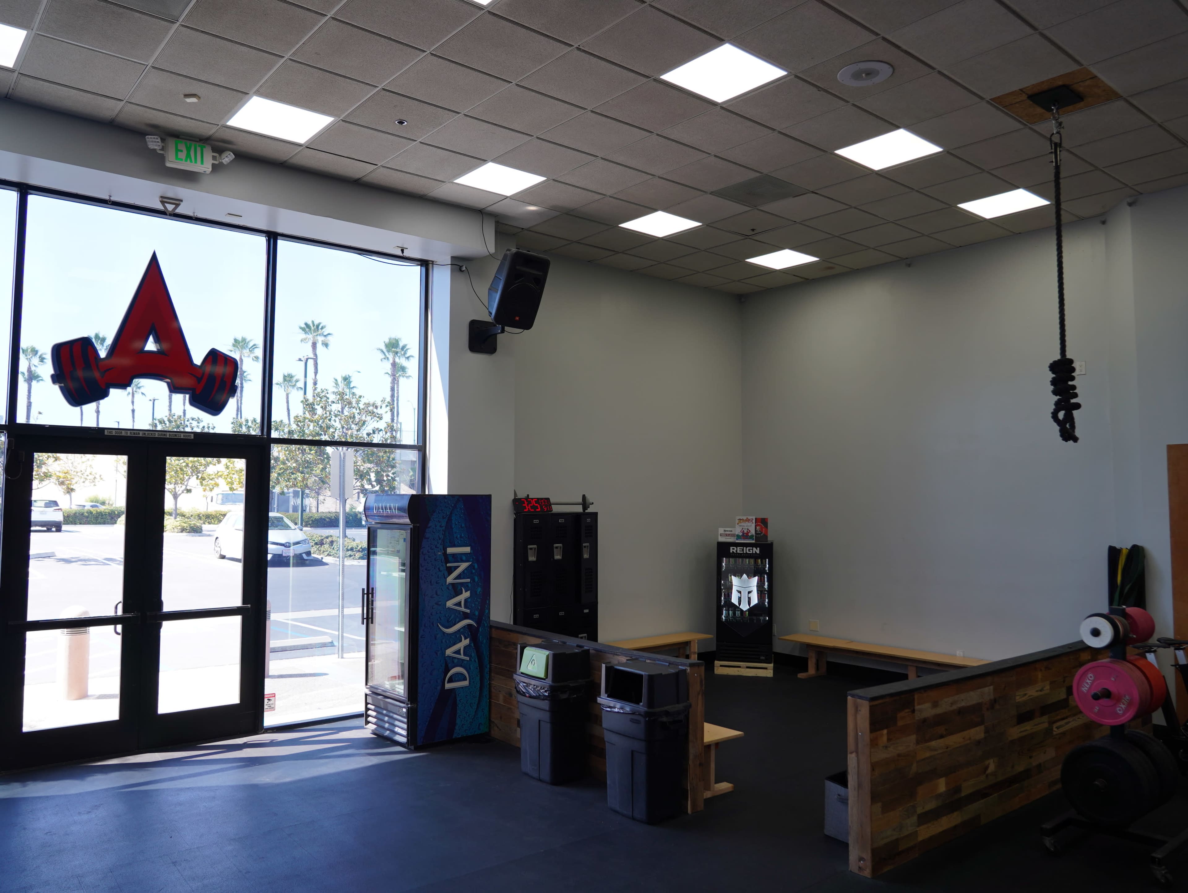 The image shows the interior of a gym with a large logo above the entrance, including racks for weights and benches along the walls.