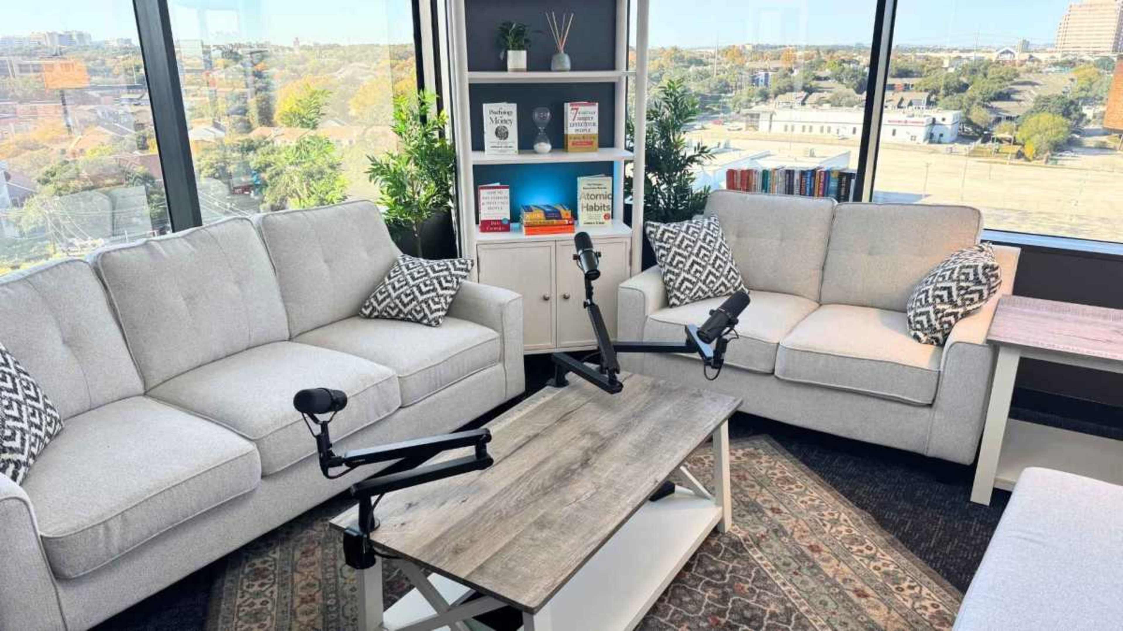 The image depicts a modern, bright lounge area featuring two light gray sofas with patterned cushions, a center coffee table, and a bookshelf filled with books, all set against a window view.