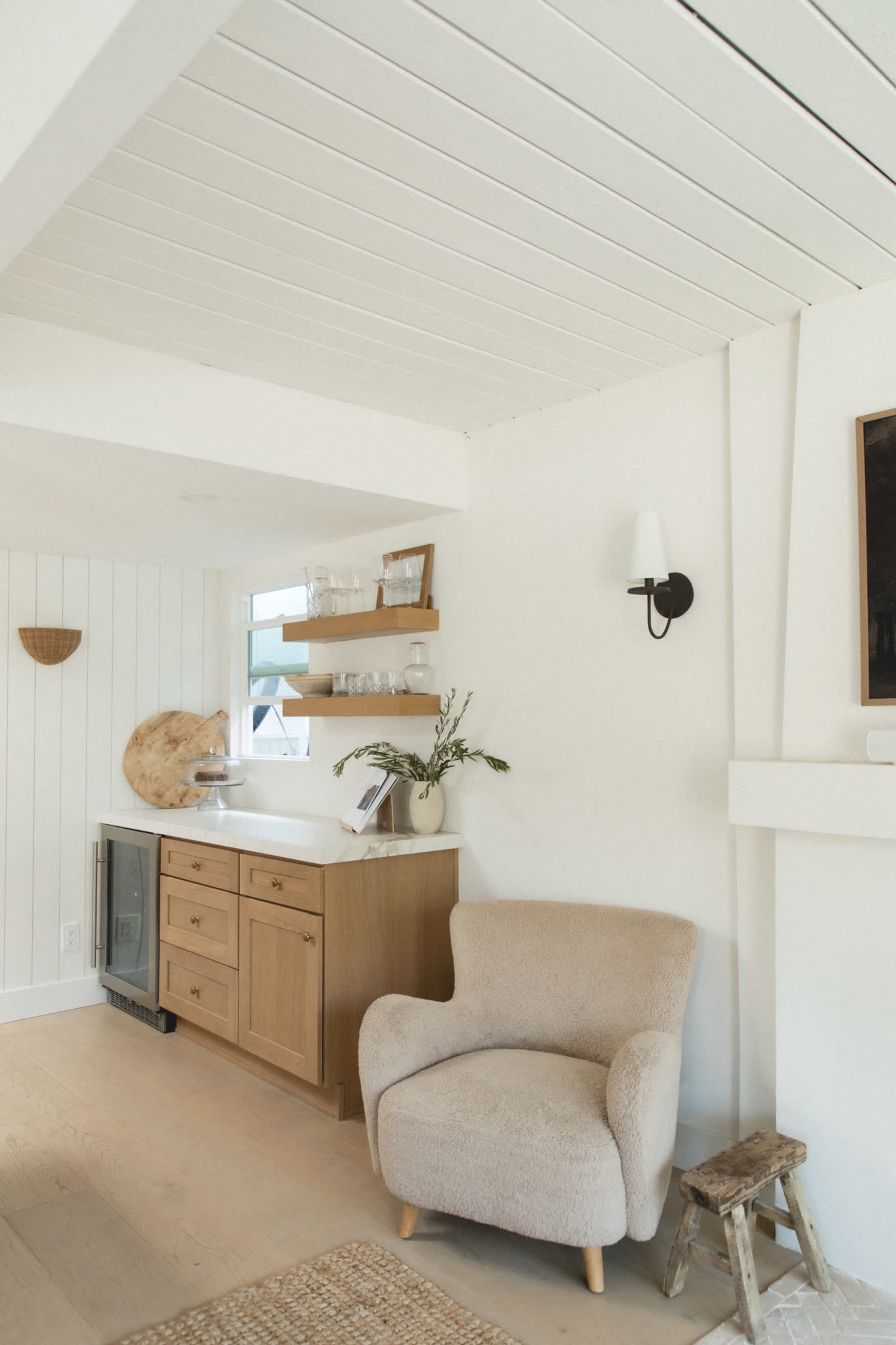The image shows a bright interior space featuring a wooden kitchen counter with shelving, a beige armchair, and light wood flooring.