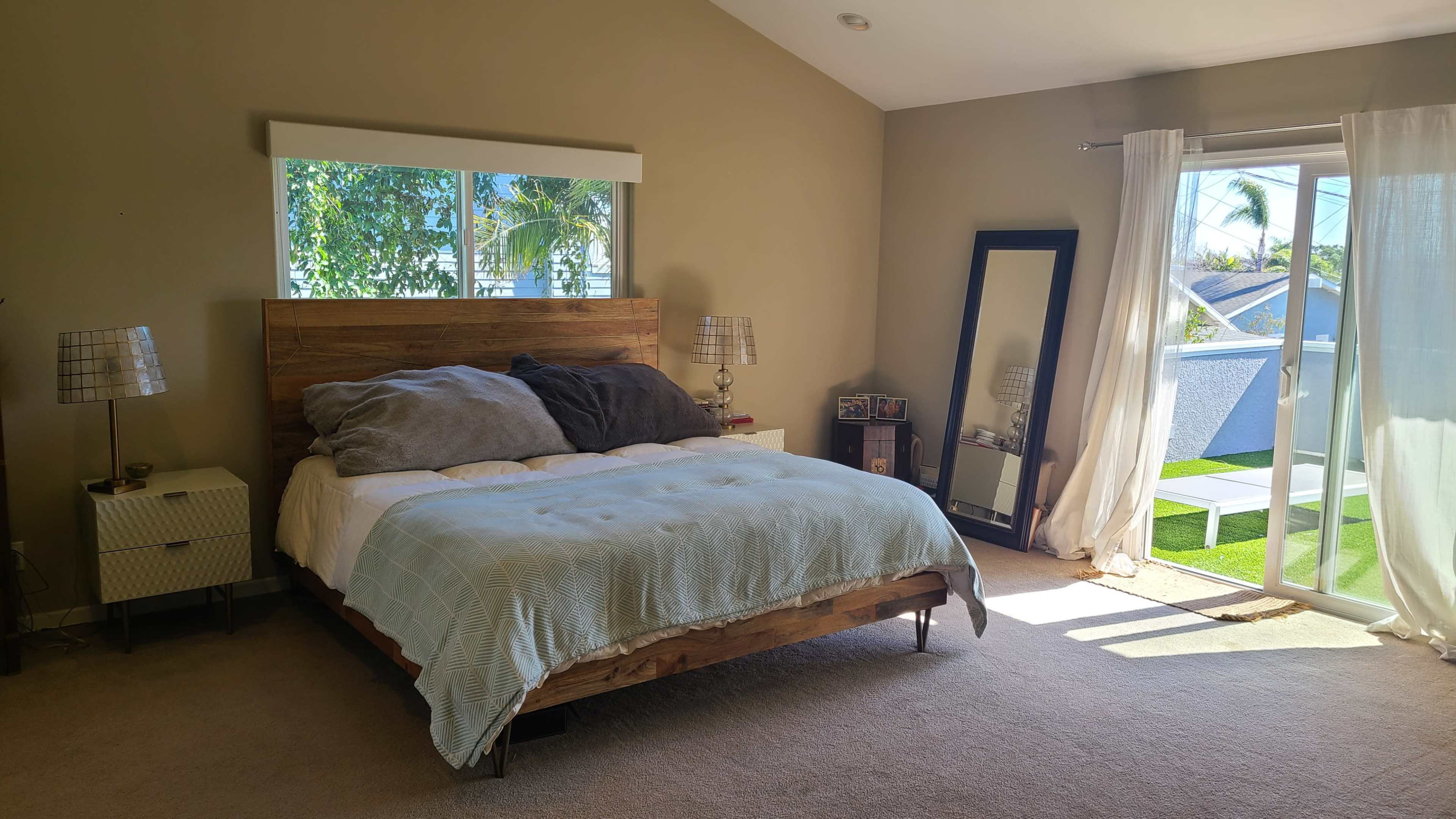 A bright bedroom featuring a wooden bed with gray bedding, a large mirror beside a window, and a sliding glass door leading to a small outdoor space.
