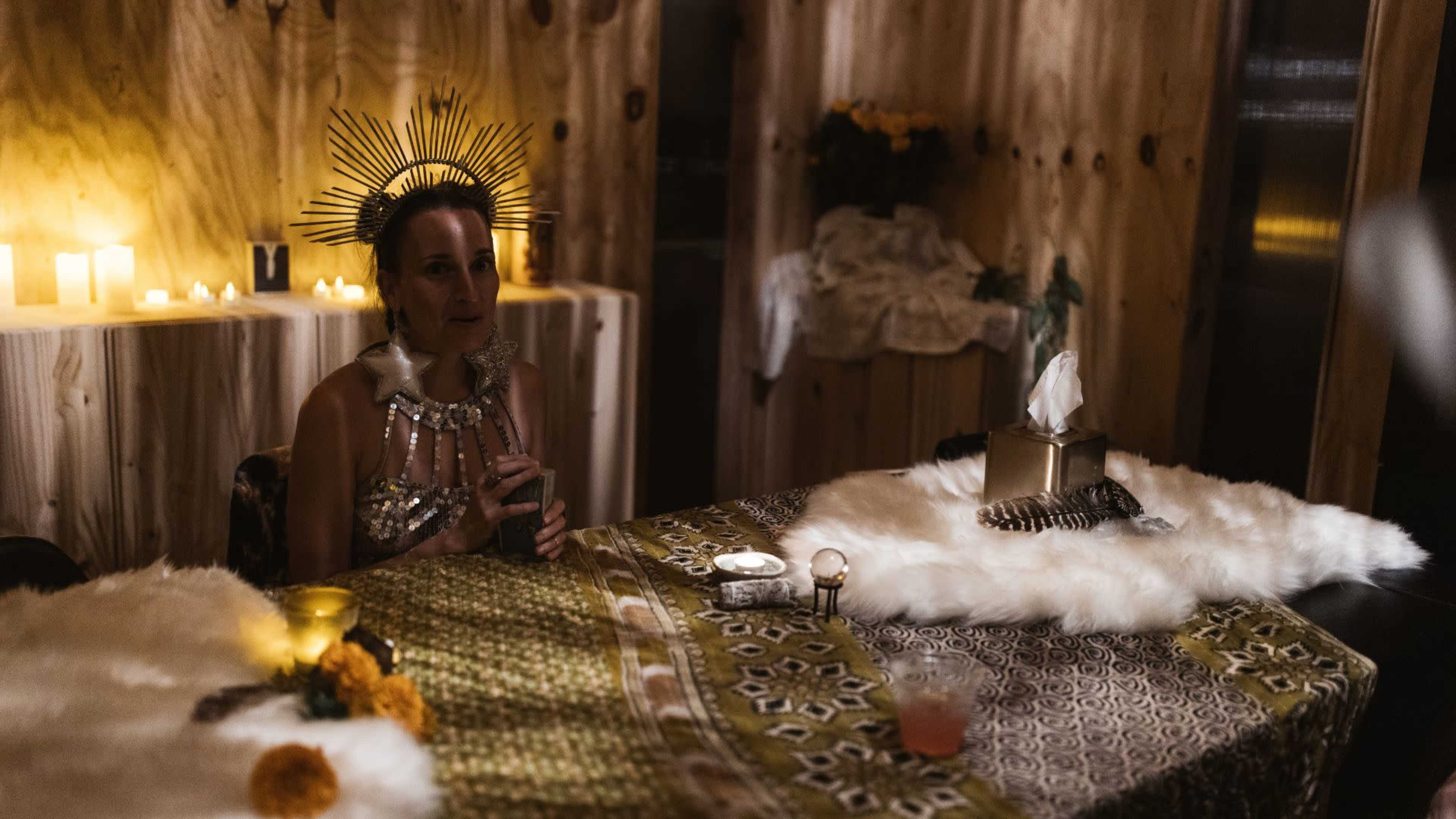 A woman wearing a decorative headpiece sits at a table adorned with candles and a floral arrangement in a dimly lit room.