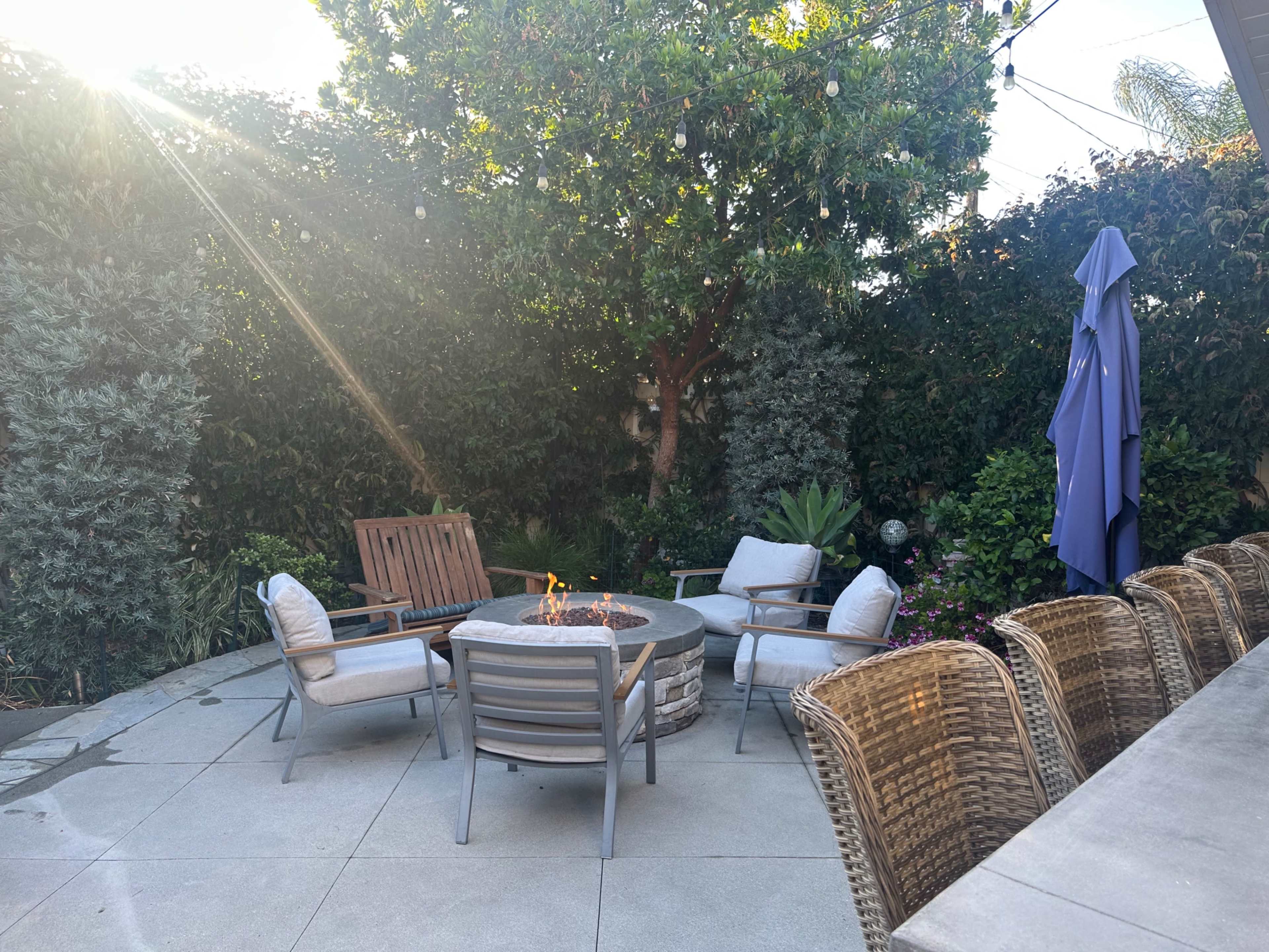 A circular patio area features a fire pit surrounded by four chairs, with lush greenery and a large umbrella nearby.