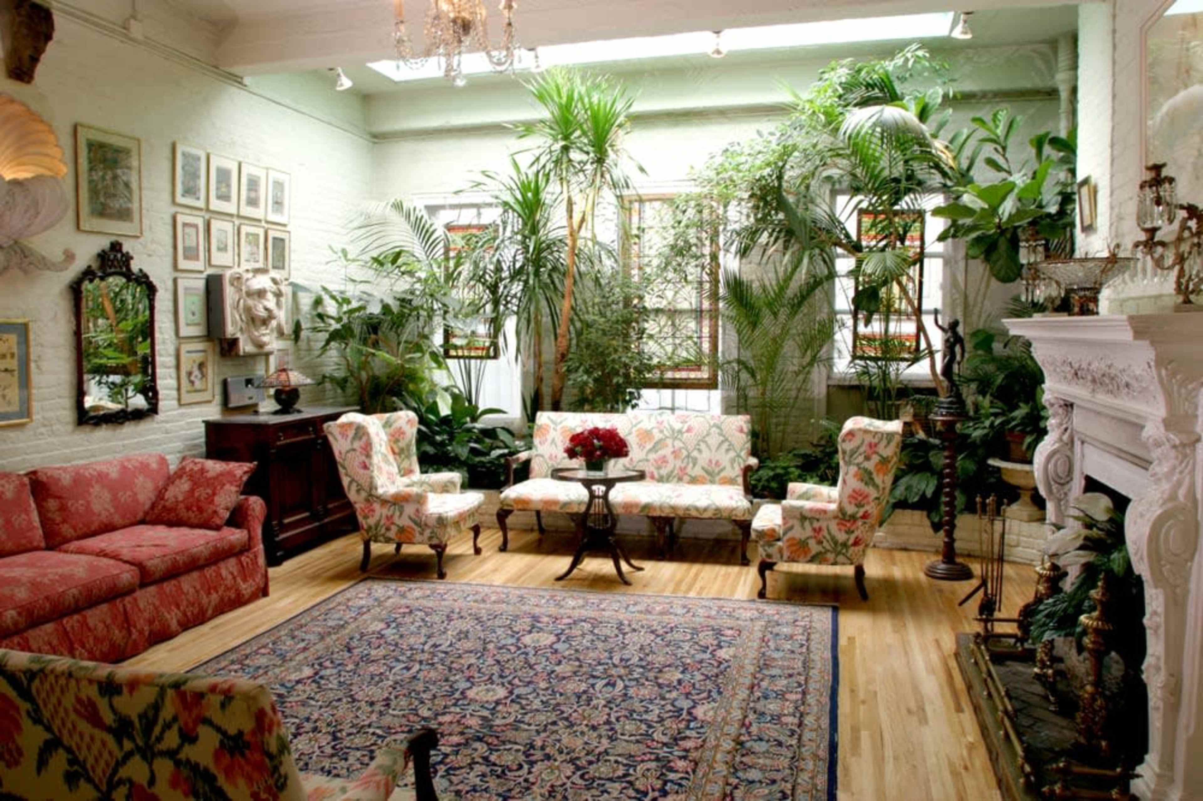 A bright living room is filled with numerous indoor plants, floral-patterned furniture, and an ornate rug.