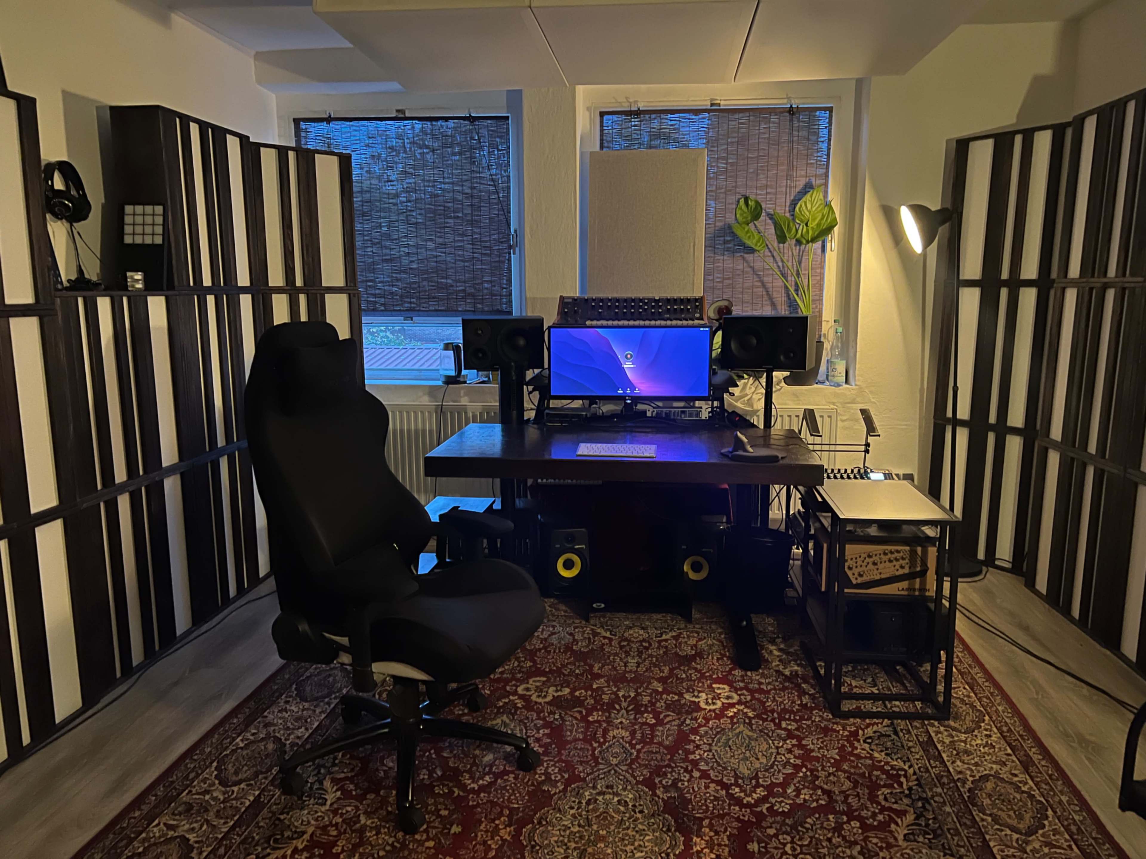 The image shows a modern workspace featuring a desk with a computer setup, flanked by sound speakers and decorative room dividers, with a chair, lamp, and a plant in a corner.