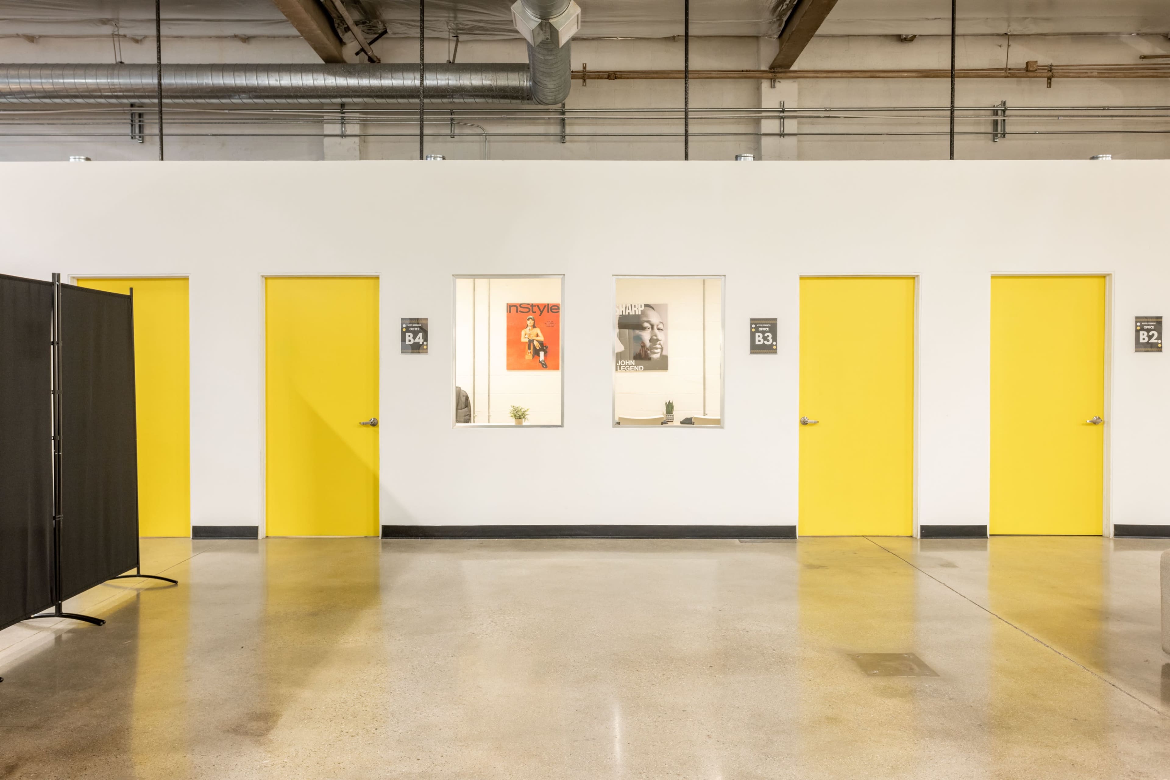 A hallway with three yellow doors and two windows displaying posters, alongside a black partition on the left.