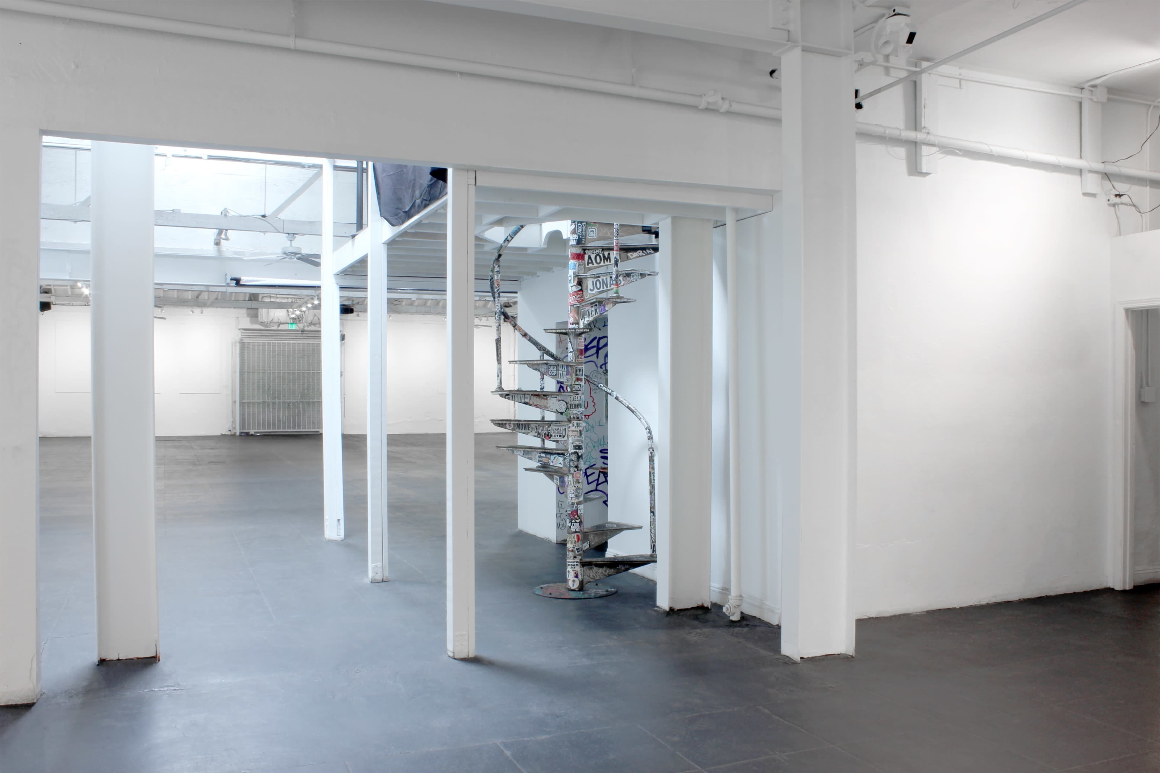 A spiral staircase, covered in stickers, stands in the center of a spacious, well-lit gallery with white walls and a dark floor.