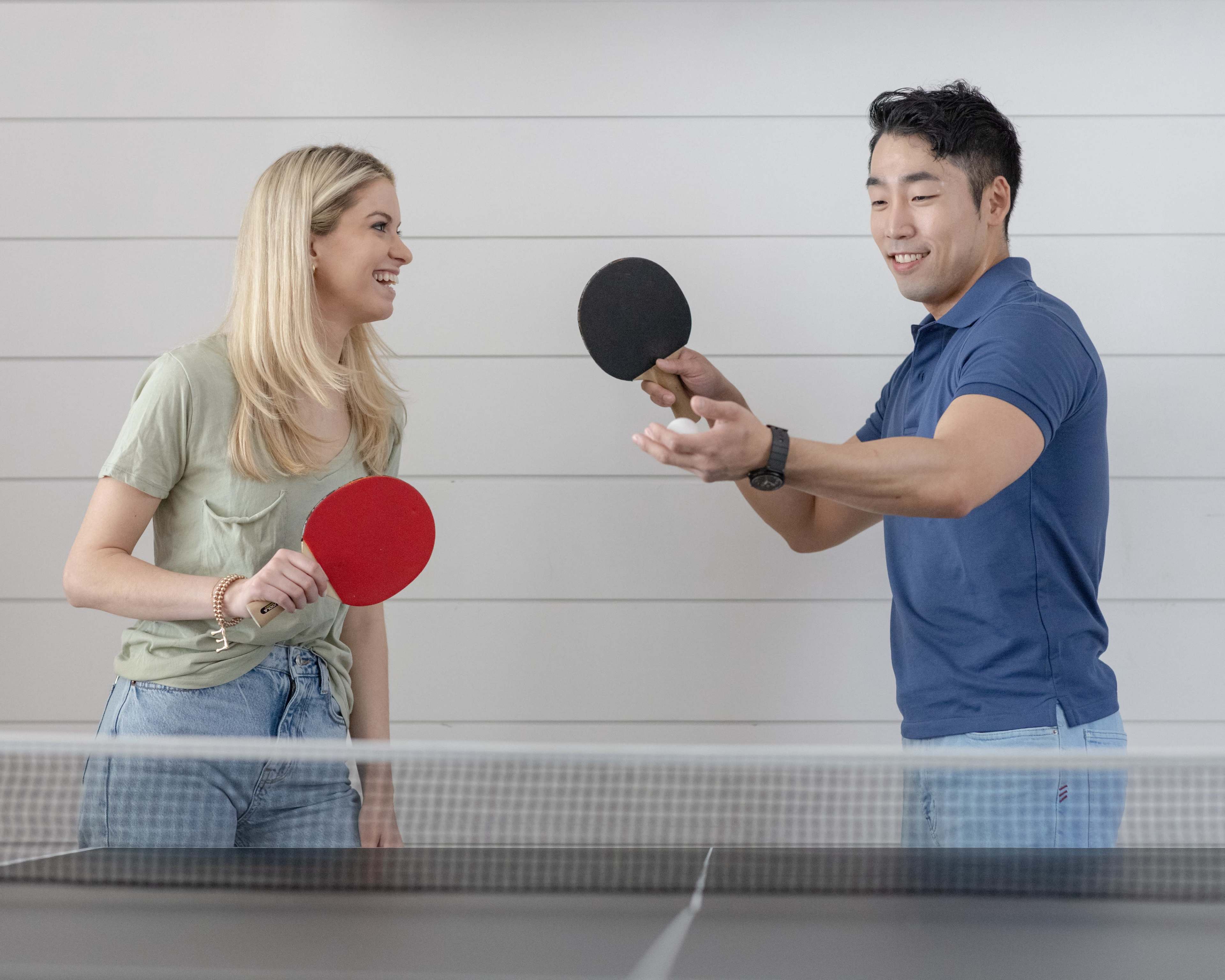 A woman and a man stand at a table tennis table, each holding a paddle and engaging in a friendly game.