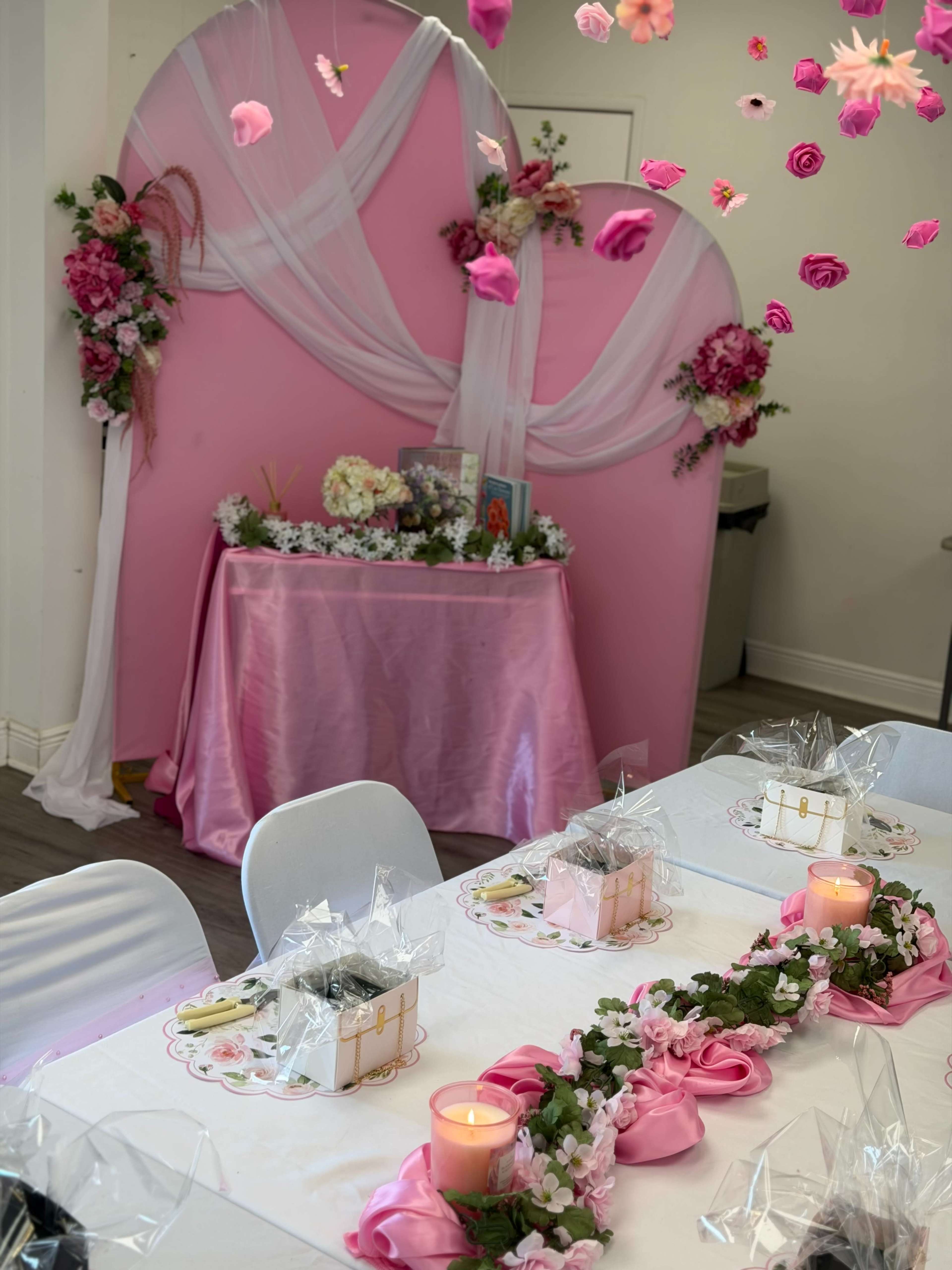 A decorated table with pink accents features floral arrangements, gift boxes, and candles, set against a pink backdrop adorned with draped fabric and flowers.