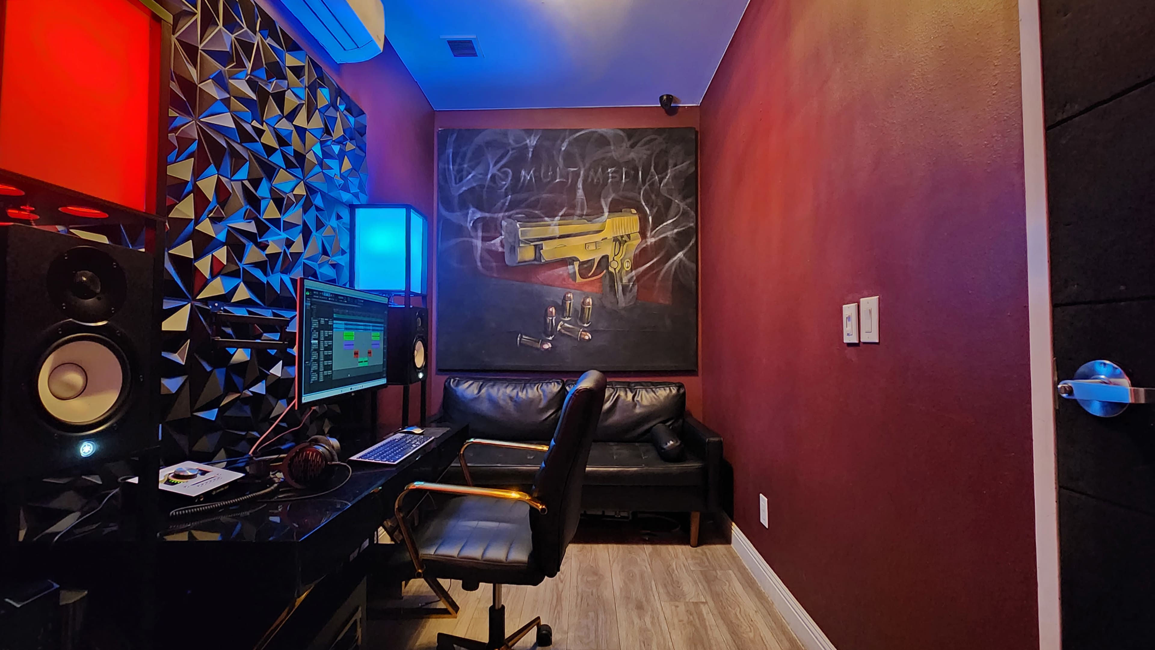 A small recording studio with a dark-red wall, a black leather sofa, and music equipment arranged on a desk.