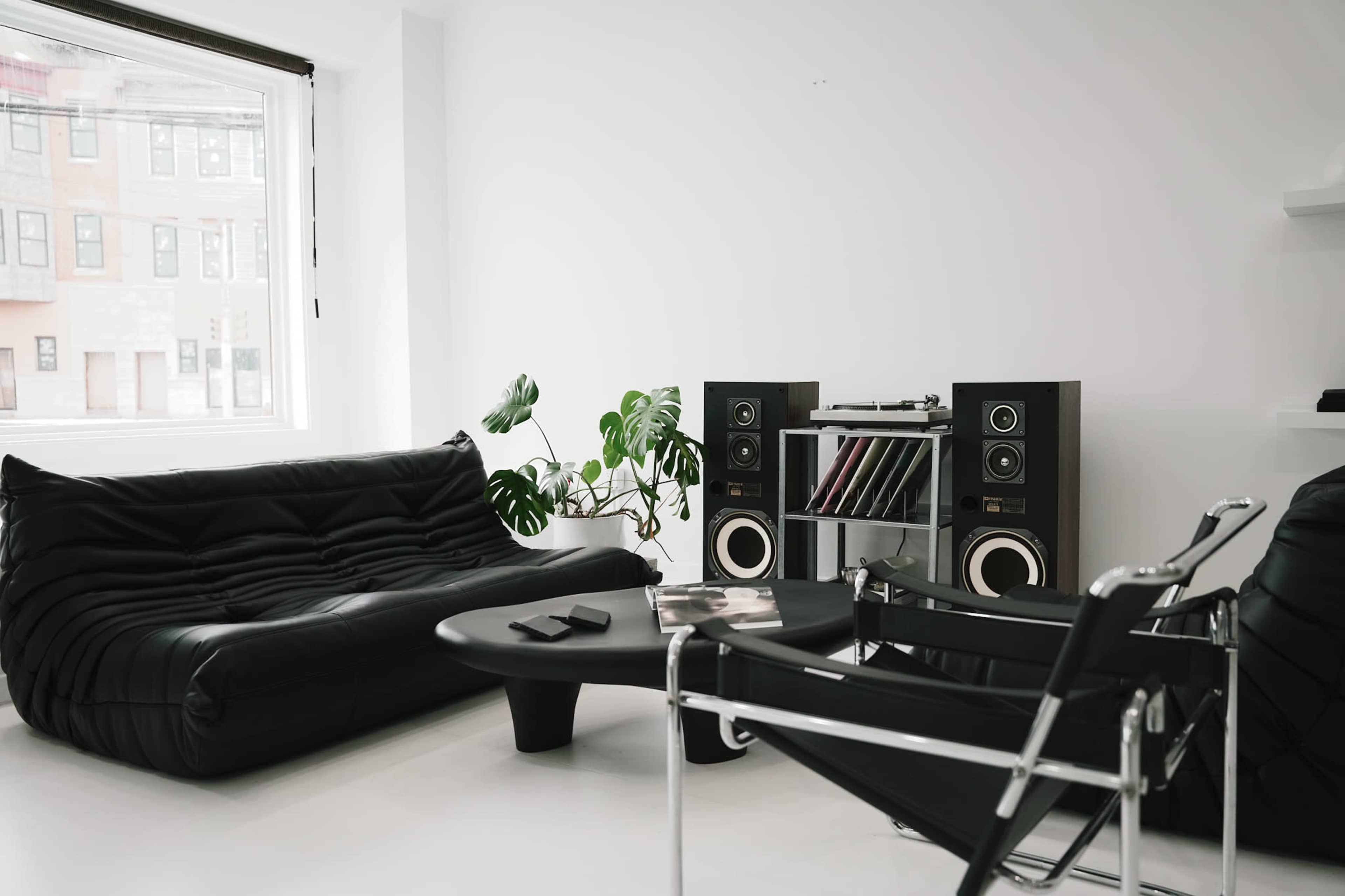 The room features a minimalist design with a black sofa, a modern chair, a round coffee table, and audio speakers on a shelf against a white wall.