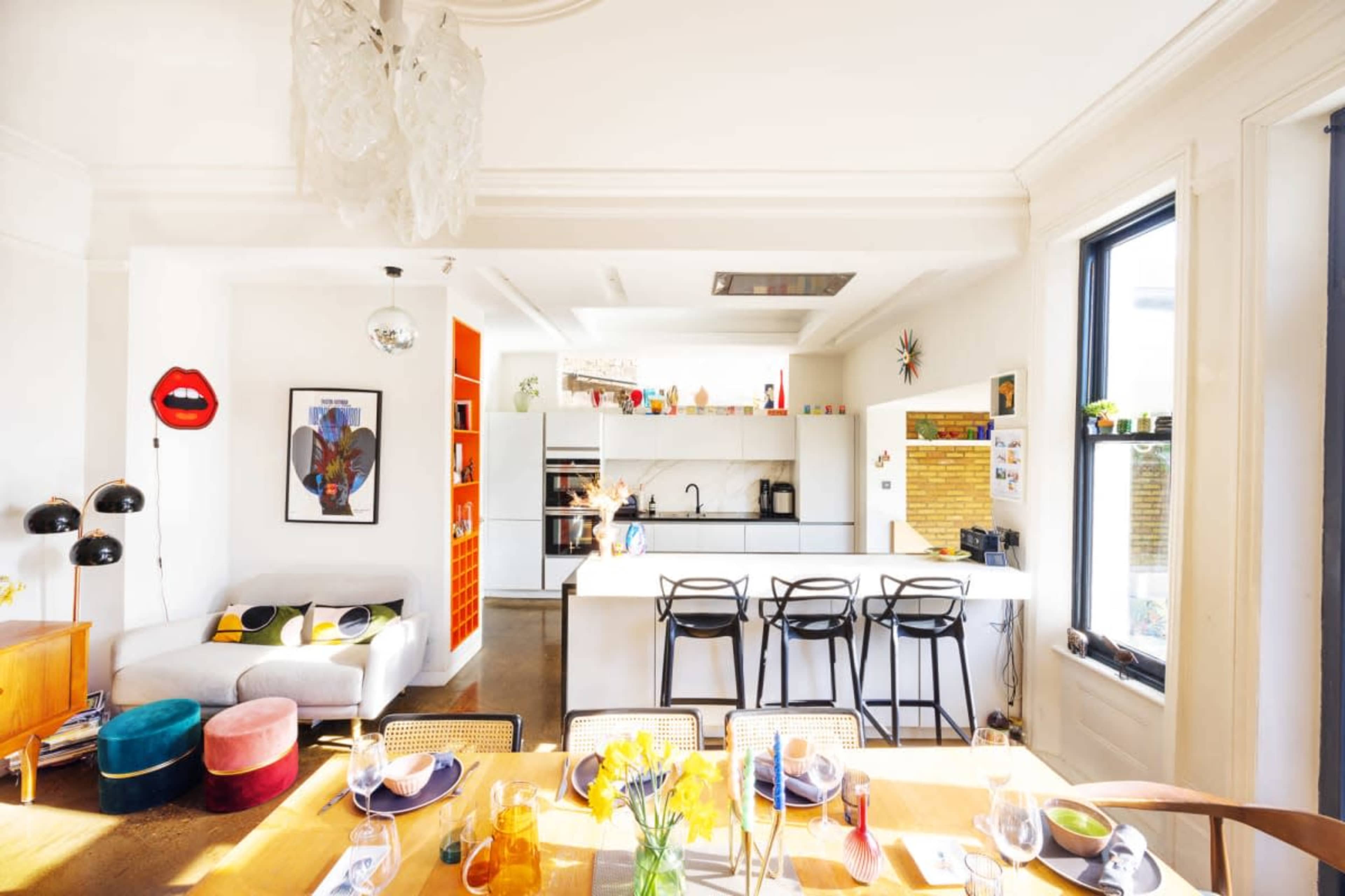 The image shows a bright, modern kitchen and dining area with a long white island, colorful chairs, and various decorative elements on the walls.