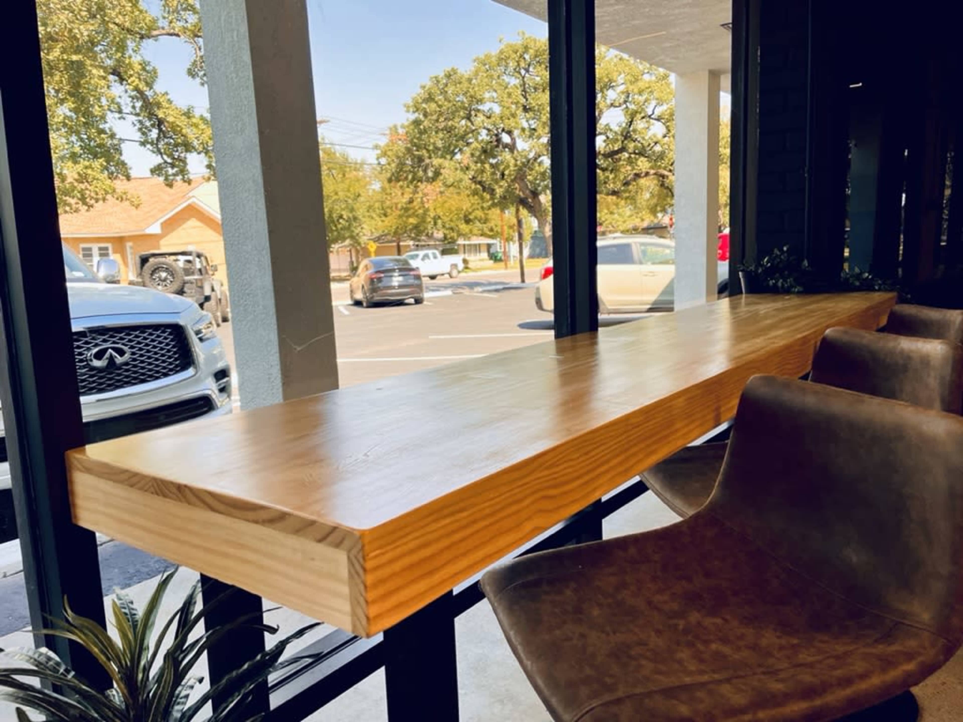 A wooden table with a long surface is positioned by a large window, overlooking a parking lot and trees outside.